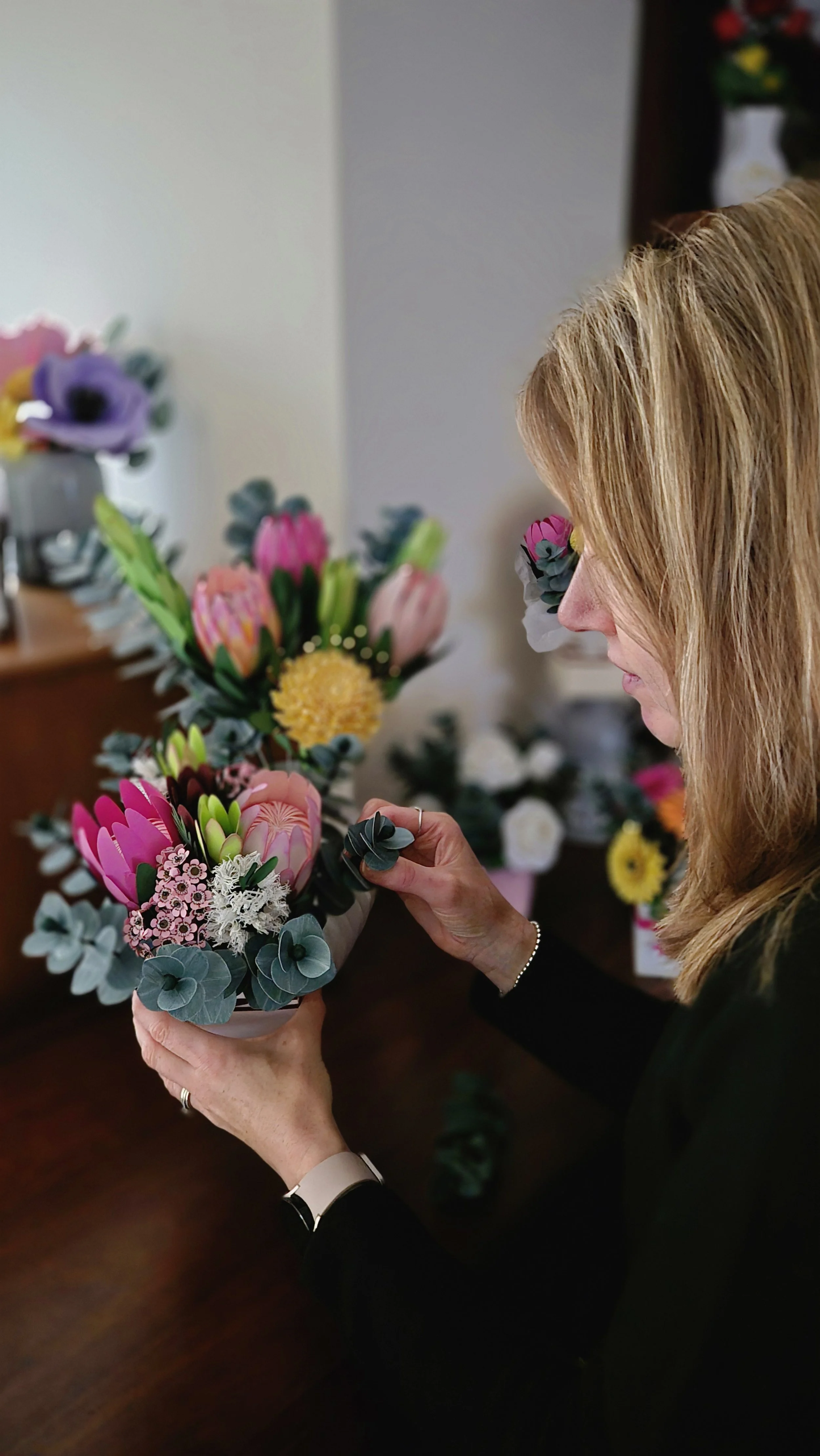 The owner Jo of Uniquely packaged arranging a colorful bouquet of flowers, including pink proteas, yellow billy balls, and other assorted blooms, in a vase on a wooden table.
