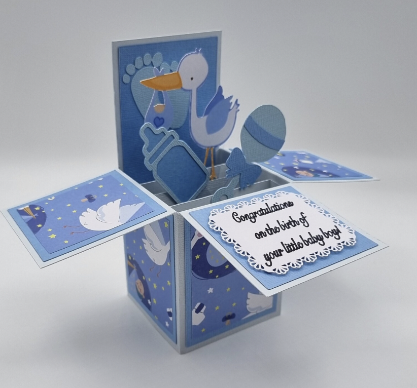 A blue pop-up greeting card with baby-themed decorations, including a stork, bottle, pacifier, and an Easter egg, with a message that says, "Congratulations on the birth of your little baby boy!"