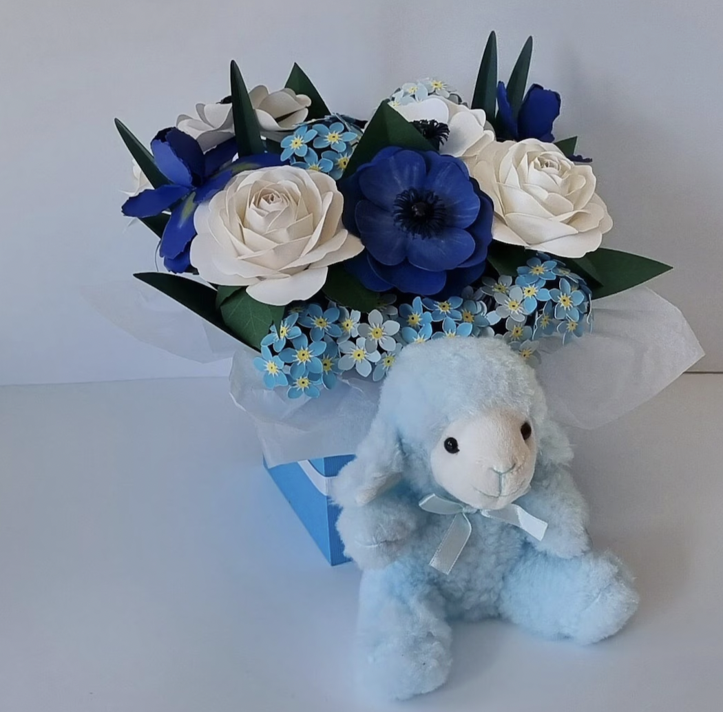 A bouquet of artificial blue and white flowers with green leaves, arranged in a box decorated with blue and white paper. In front of the bouquet is a small plush white bunny with a satin ribbon around its neck.