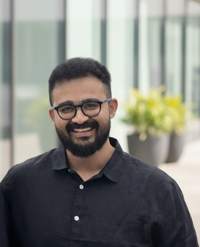 A smiling man with glasses and a beard wearing a black shirt, standing indoors with large windows and potted plants in the background.