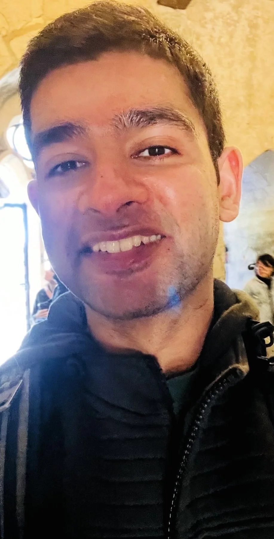 Close-up of a young man with short dark hair smiling, wearing a black jacket, indoors with a warm, brightly lit background and other people in the distance.