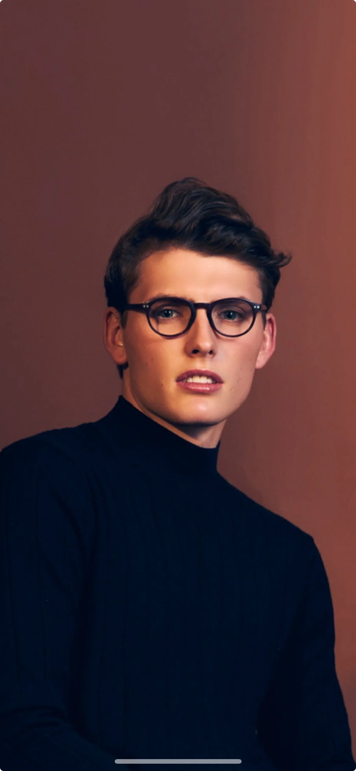 A young woman with short, dark hair, wearing round black glasses and a black turtleneck, looking at the camera against a plain brown background.