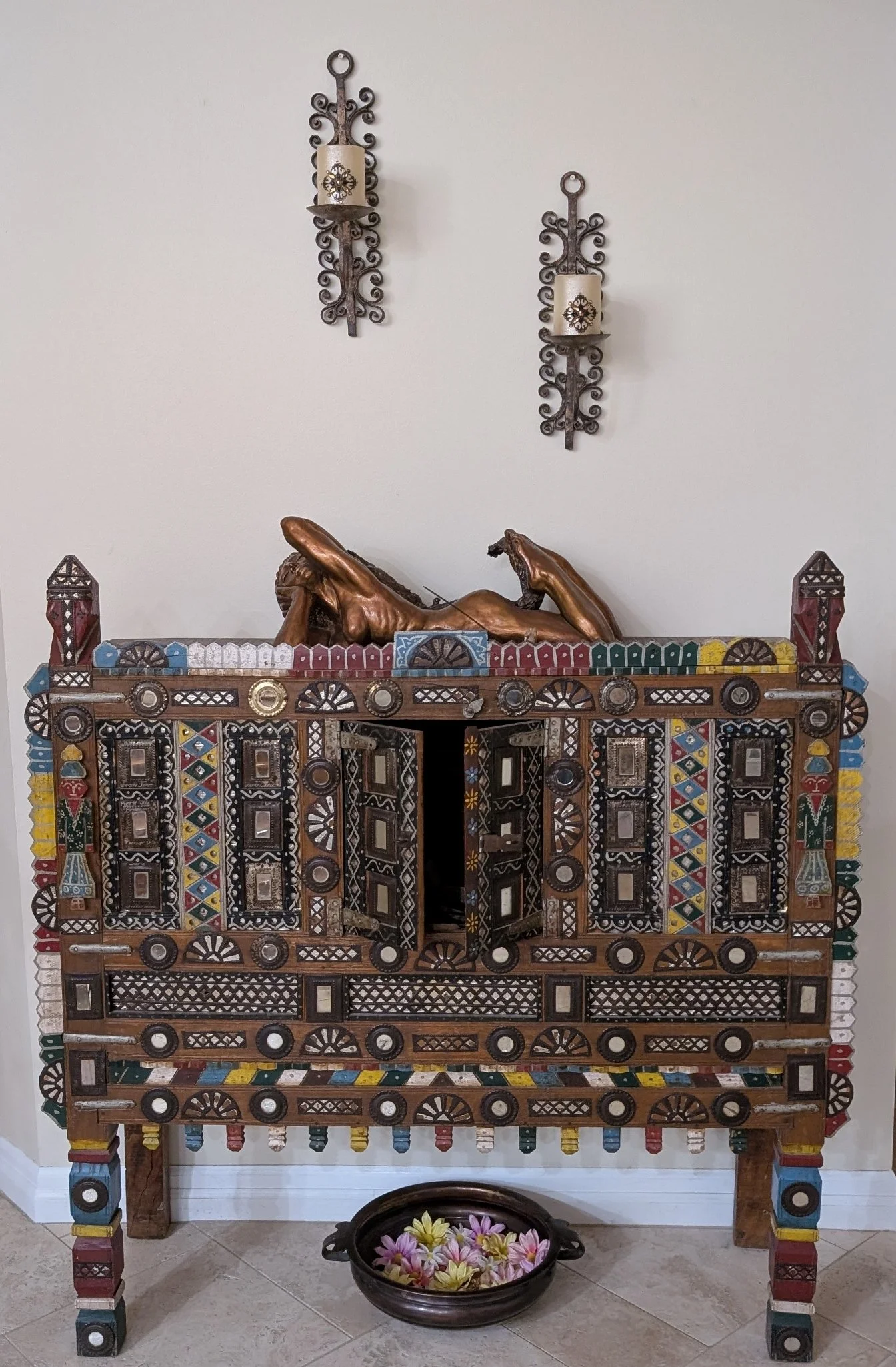 A colorfully painted Indian damchiya or dowry chest, a pair of wrought iron wall candles sticks above it, and a brown metal urli or bowl below filled with faux "floating" flowers