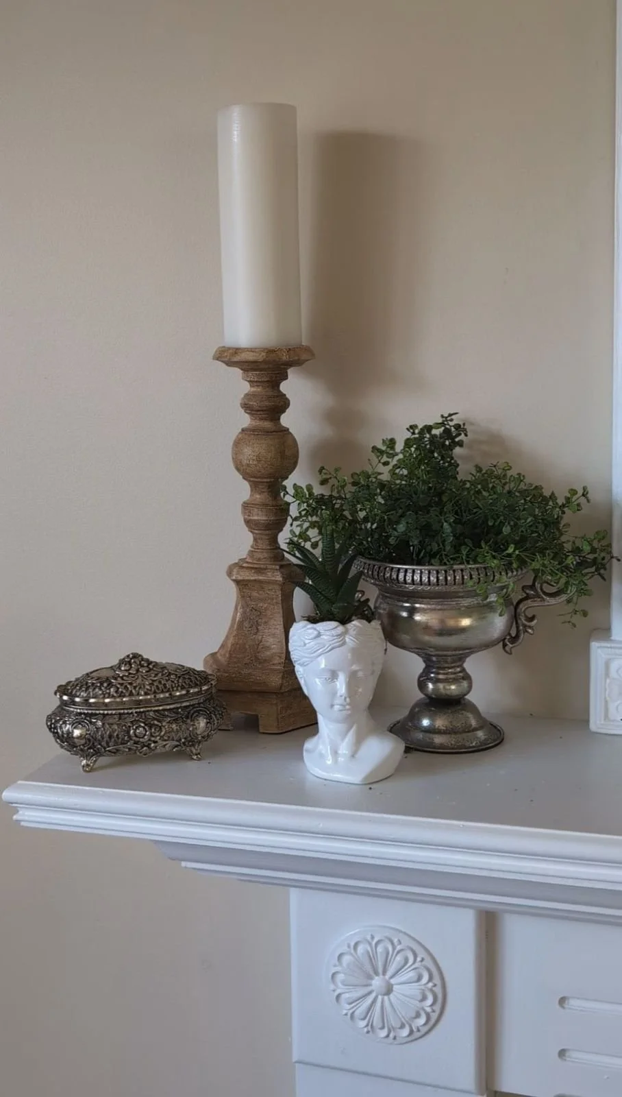 A mantle top with two silver vessels, a yellow-brown oak candlestick and a white bust planter containing a succulent