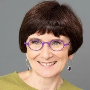 A woman with short brown hair, purple glasses, and hoop earrings smiling at the camera, wearing a green top against a gray background.