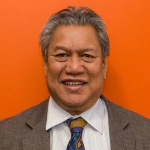A smiling man with gray hair wearing a suit and tie against an orange background.