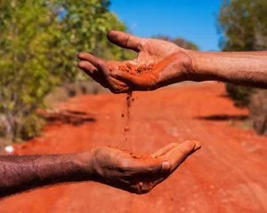 Two hands, one older and one younger, with dirt falling from the outstretched finger of the older hand towards the younger hand against a background of red soil and trees.