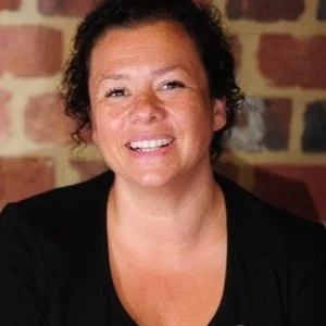 A smiling woman with curly hair wearing a black top, standing in front of a brick wall.