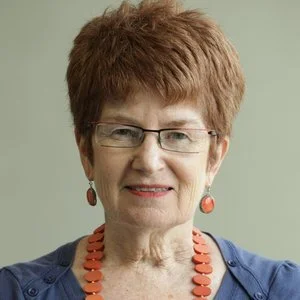 A mature woman with short reddish-brown hair, glasses, and earrings, smiling at the camera against a plain light green background.