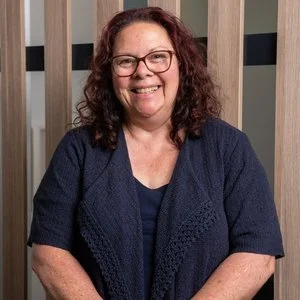 A woman with curly hair, glasses, and a navy blue top, smiling in front of a wooden panel background.