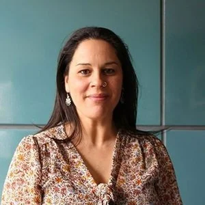A woman with long dark hair, wearing earrings and a patterned blouse, standing in front of a teal-colored wall or cabinet.