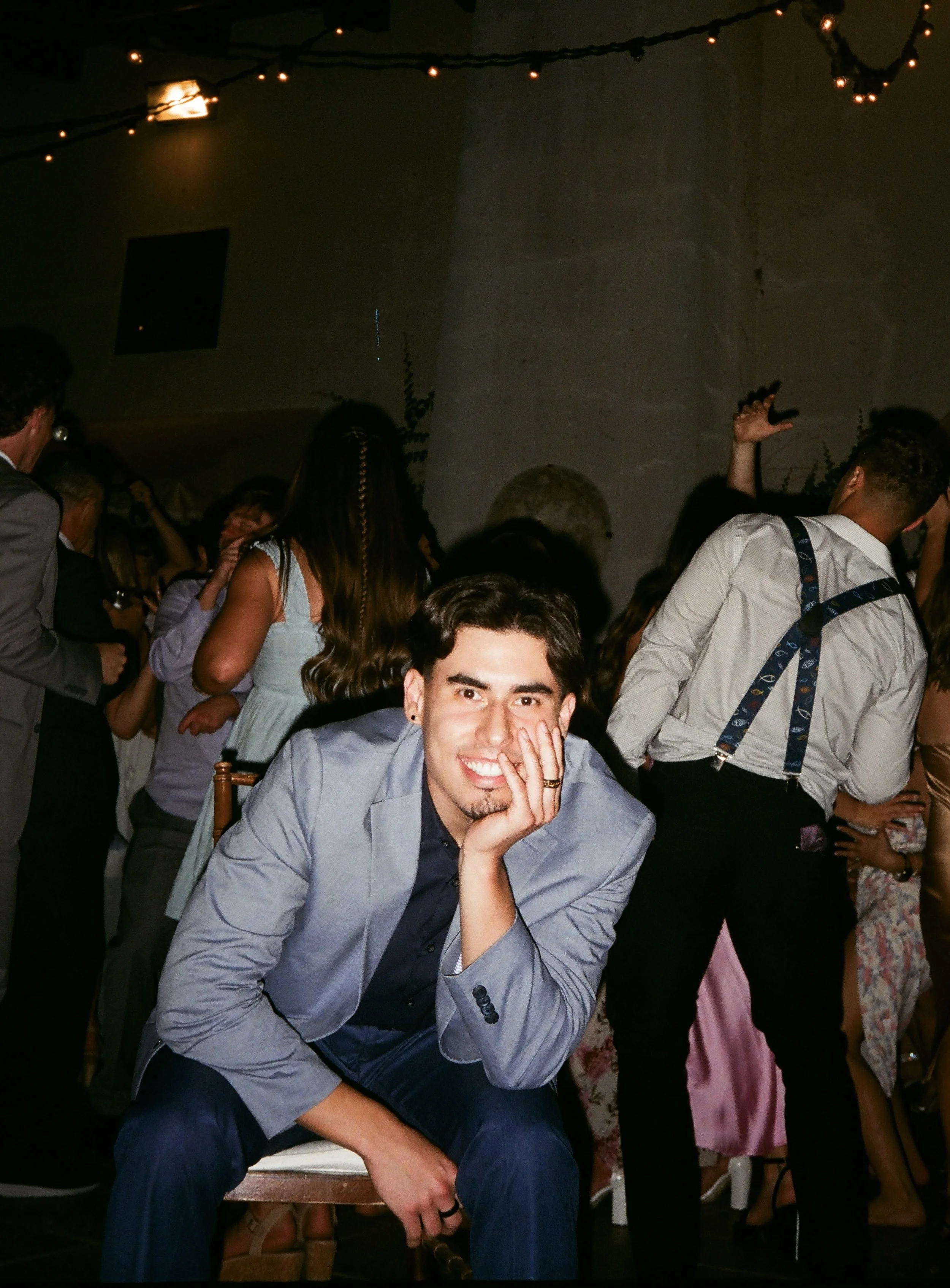 A group of people at a party, with the focus on a young man sitting in front, smiling and resting his face on his hand. He is dressed in a light gray blazer and dark shirt. The background shows other people socializing, some with drinks, and a dark wall with string lights hanging overhead.