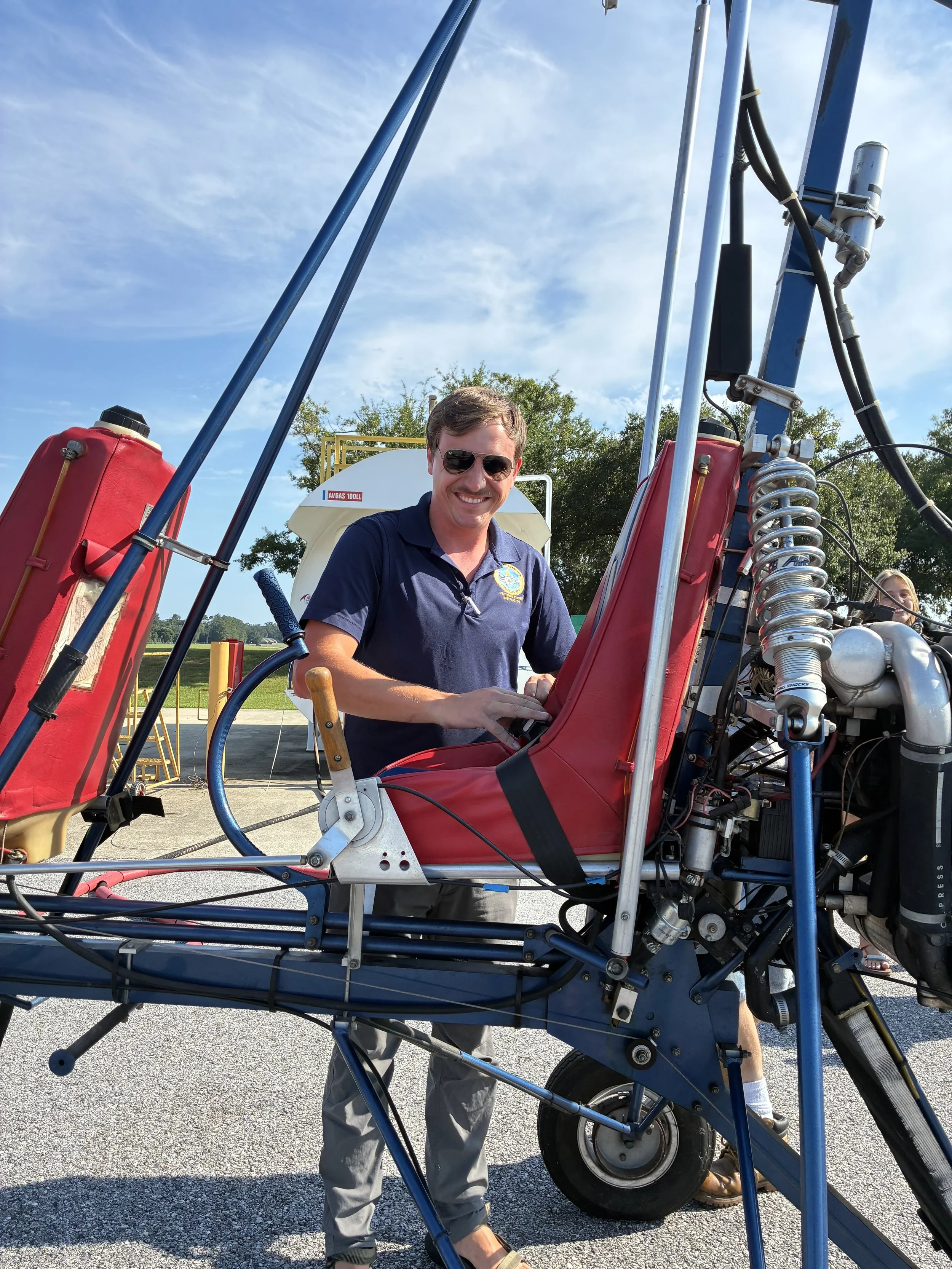 A man smiling and working on a small aircraft or ultralight vehicle outdoors on a clear day, with trees and a cloudy sky in the background.