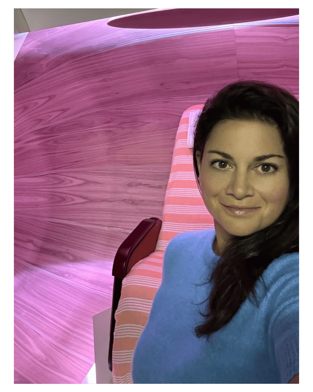 Selfie of a woman with dark hair smiling, wearing a blue top, in front of a large pink and purple abstract artwork and striped pink and white furniture.