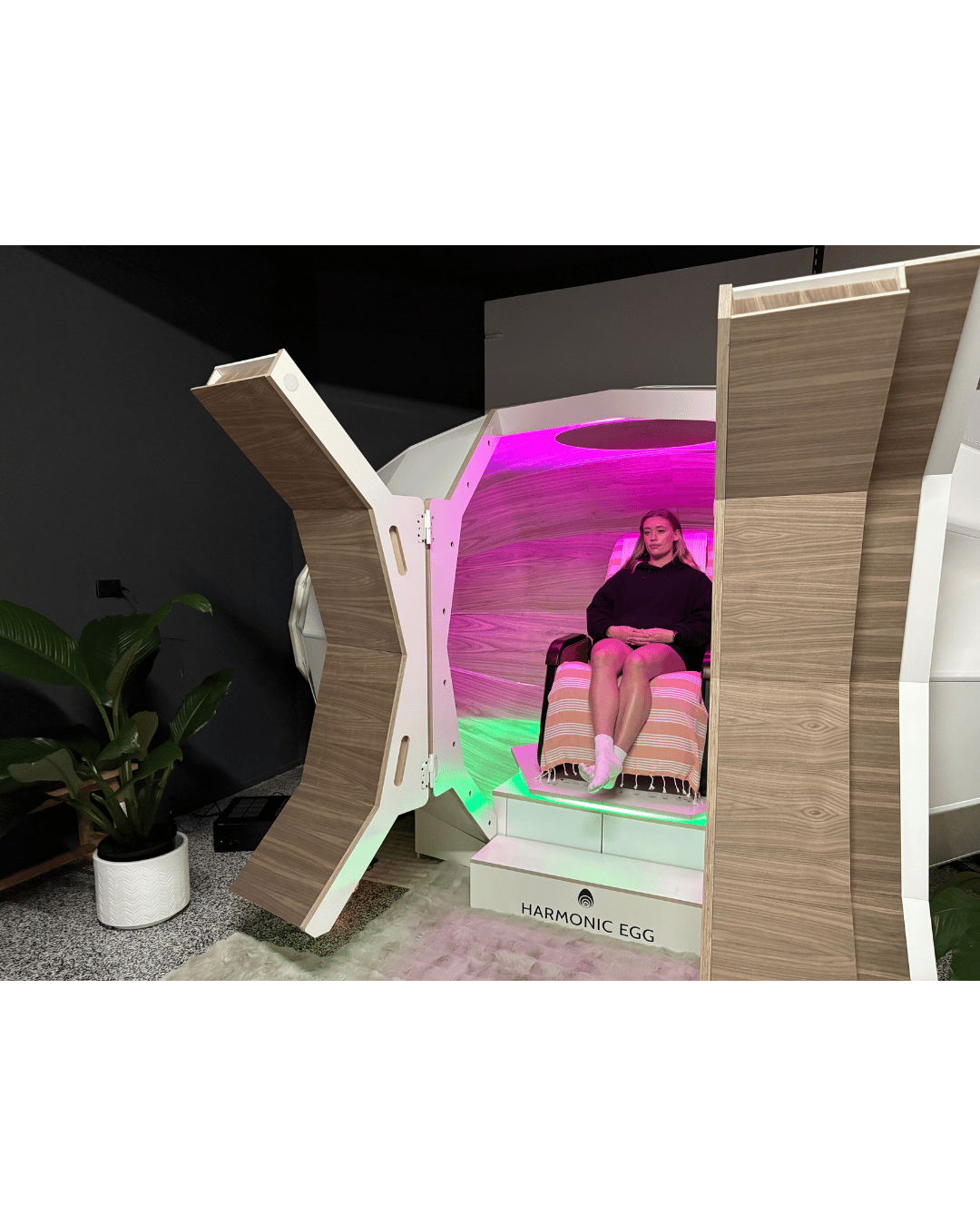 A woman sitting inside a futuristic device labeled 'Harmonic Egg,' with pink and green lighting, surrounded by modern wooden and white panels and a green potted plant nearby.