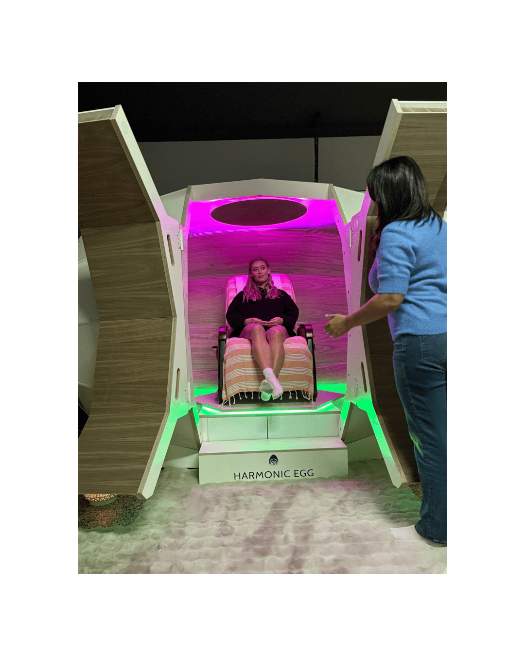 A woman sitting in a pod-like relaxation chamber with pink and green LED lighting, with an attendant standing nearby.