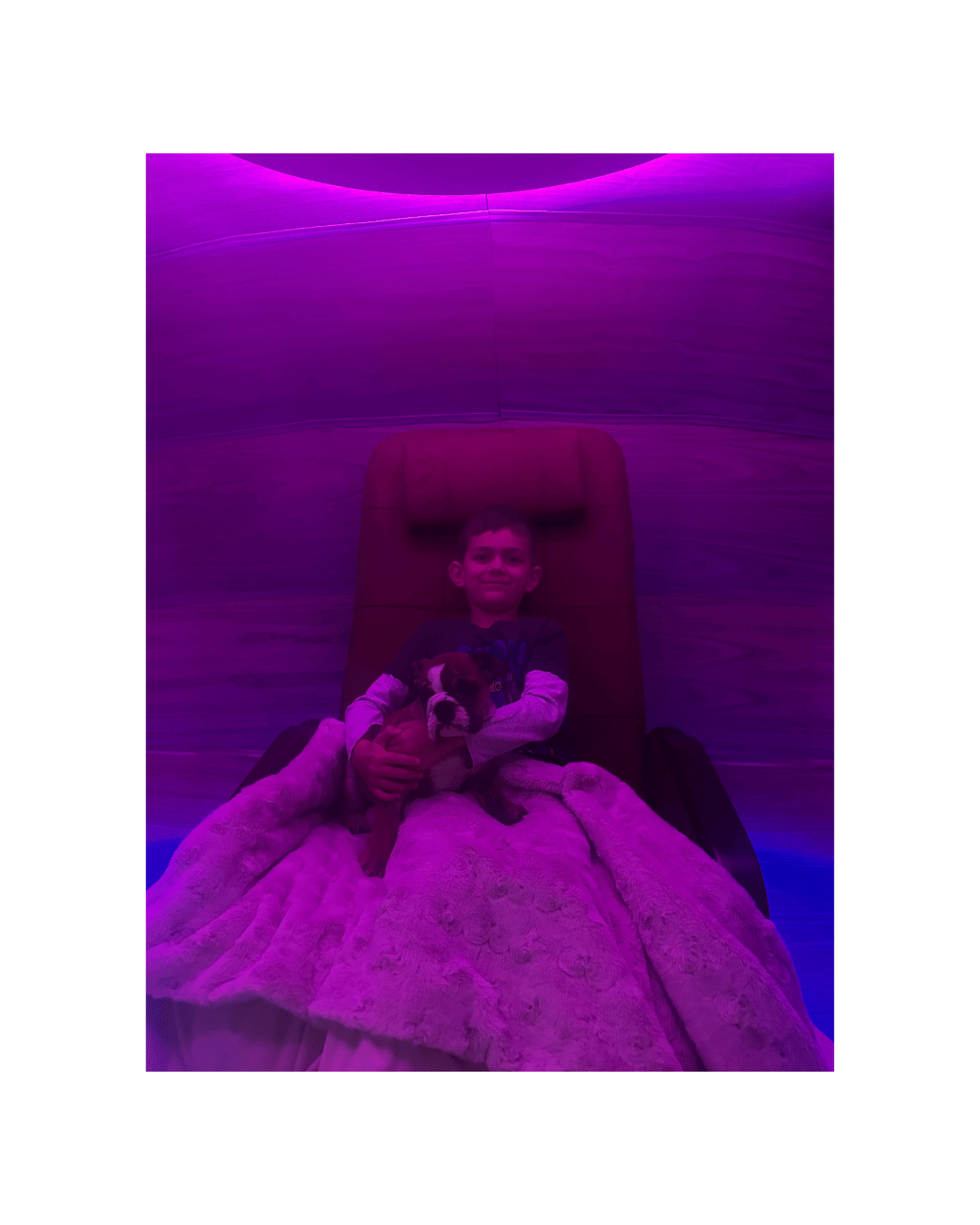 A young boy smiling while sitting in a lounge chair, holding a puppy, under purple and pink neon lighting.