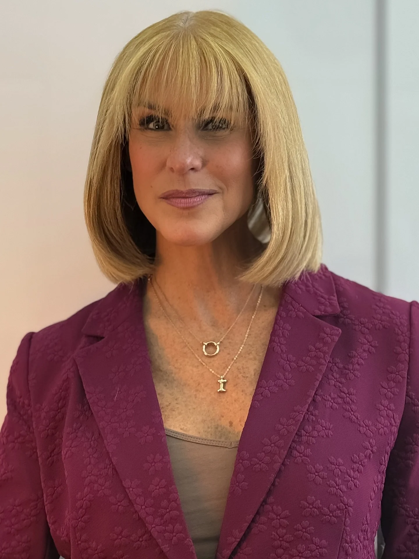 Close-up of a woman with blonde hair and bangs wearing a purple textured blazer and layered necklaces, standing indoors with a plain background.