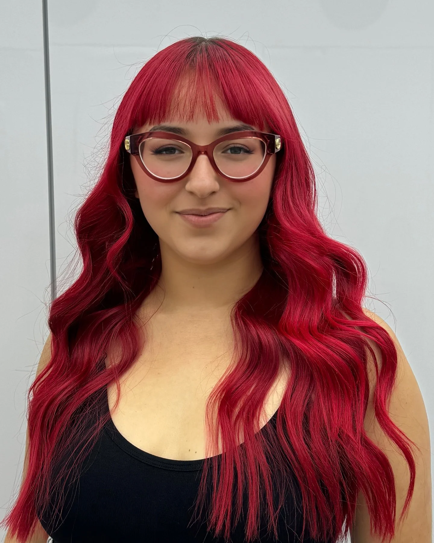 A woman with long, wavy red hair, wearing large red glasses, smiling at the camera against a plain light gray background.