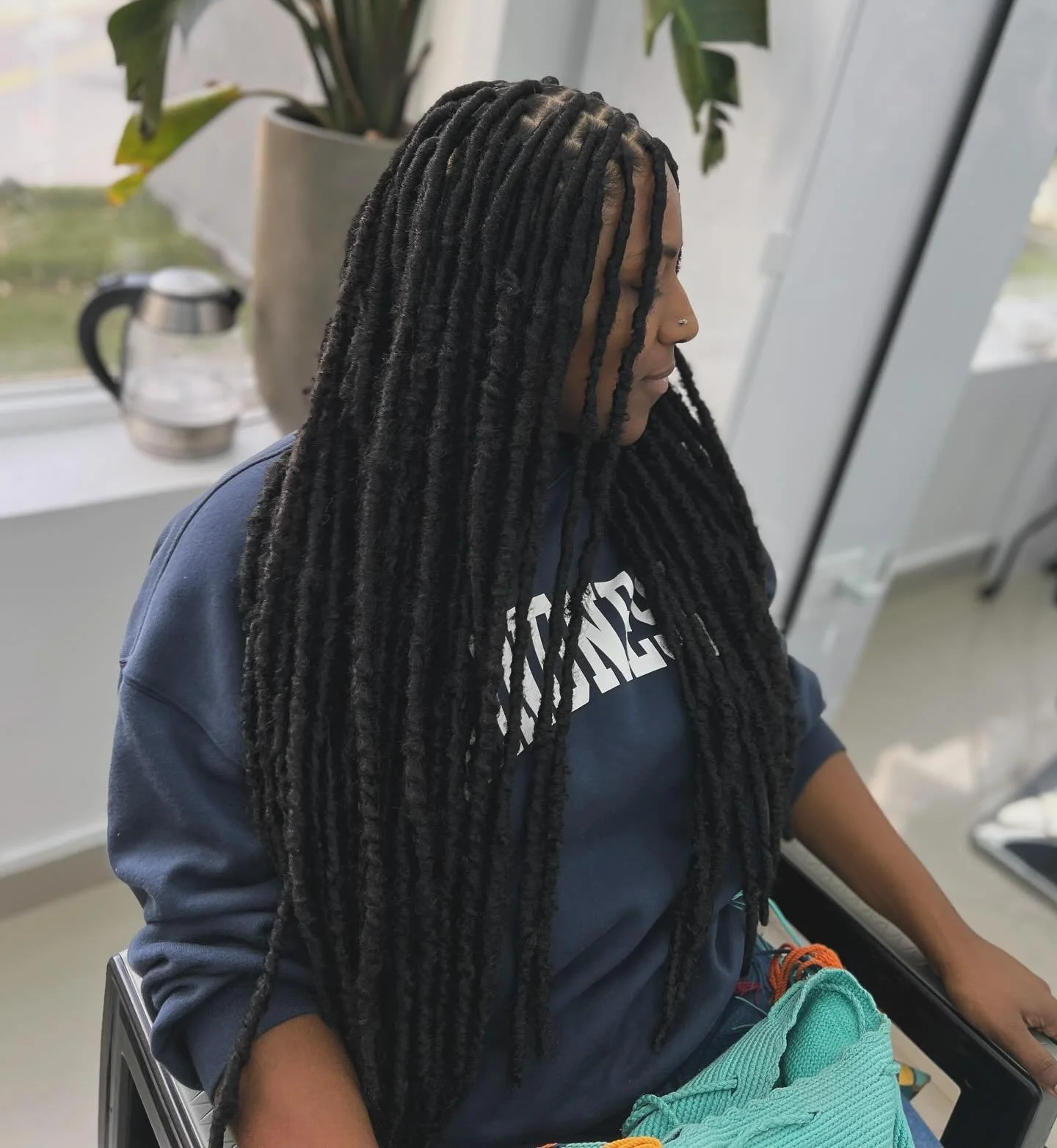 Person with long dreadlocks sitting in a room, wearing a navy blue hoodie with partially visible white text, holding a teal cloth, with a large potted plant and a window in the background.