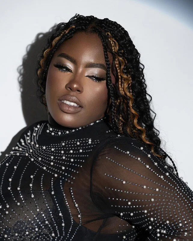 A woman with curly, dark hair with blonde highlights, posing against a gray and white background. She is winking with one eye and wearing a black, sheer, rhinestone-embellished top.