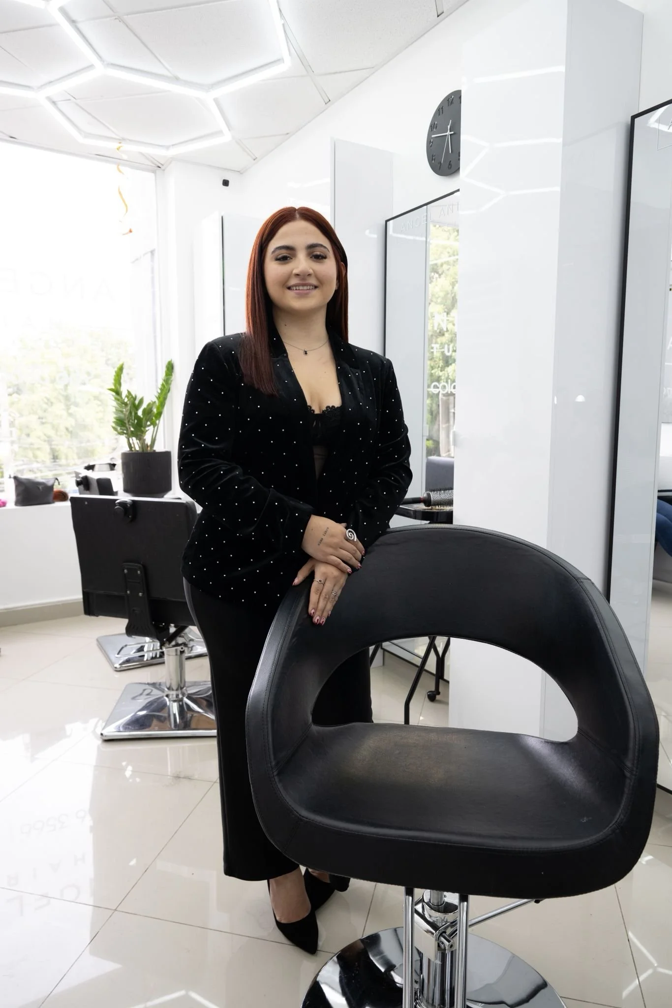 A woman with red hair in a professional black suit stands in a modern, bright salon or spa, smiling. The salon has white walls, a stylish ceiling, a clock on the wall, and black salon chairs, with sunlight coming through large windows.