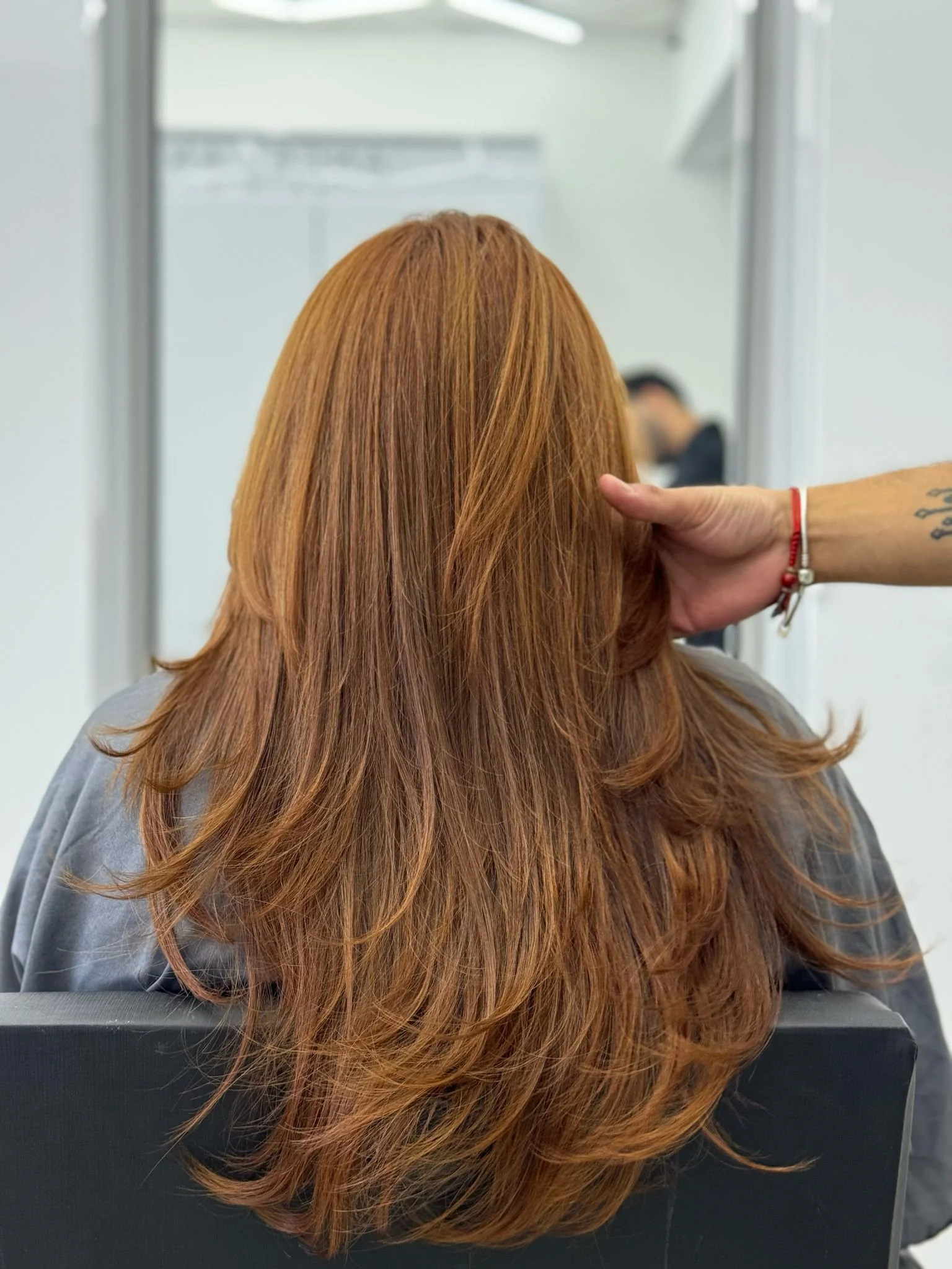 Back of a person's head with long, wavy, reddish-brown hair, seated in front of a mirror in a salon.