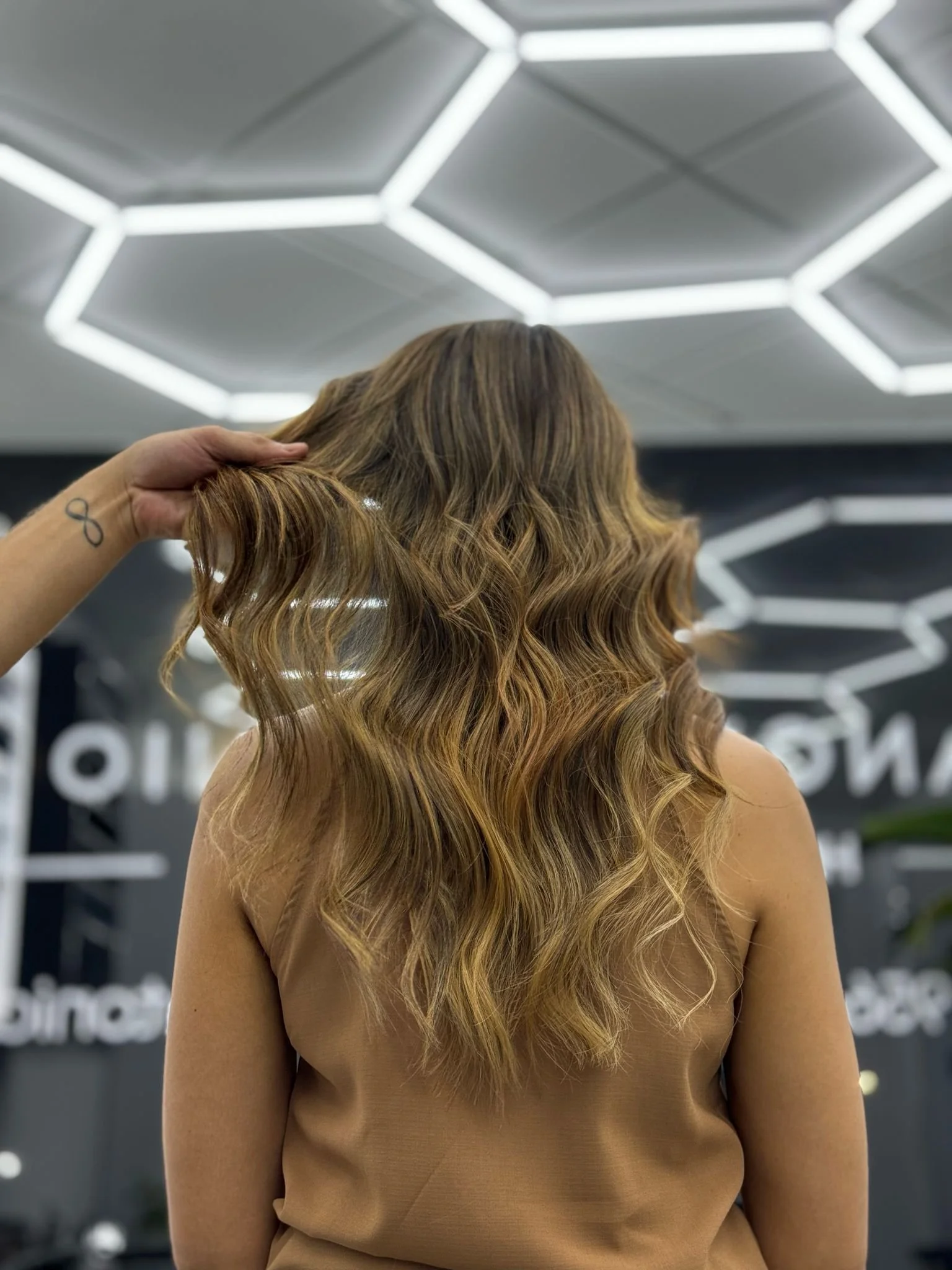 A woman with long, wavy, light brown hair, wearing a tan sleeveless top, is touching her hair with her left hand. The background appears to be an indoor space with geometric lighting on the ceiling and partially visible white text.
