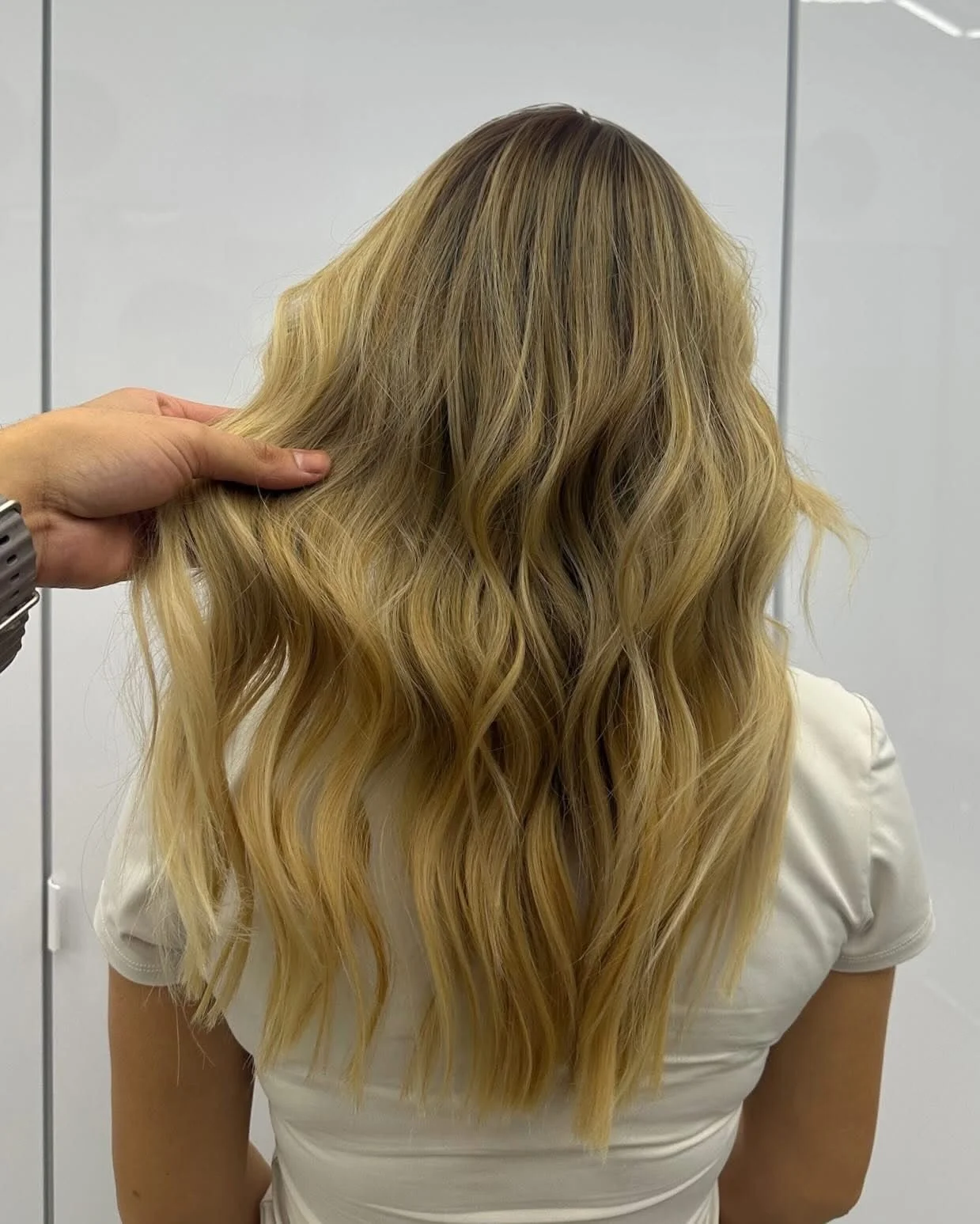 A person with long, wavy blonde hair, wearing a white shirt, is being touched on the hair by a hand near the left side of the image.