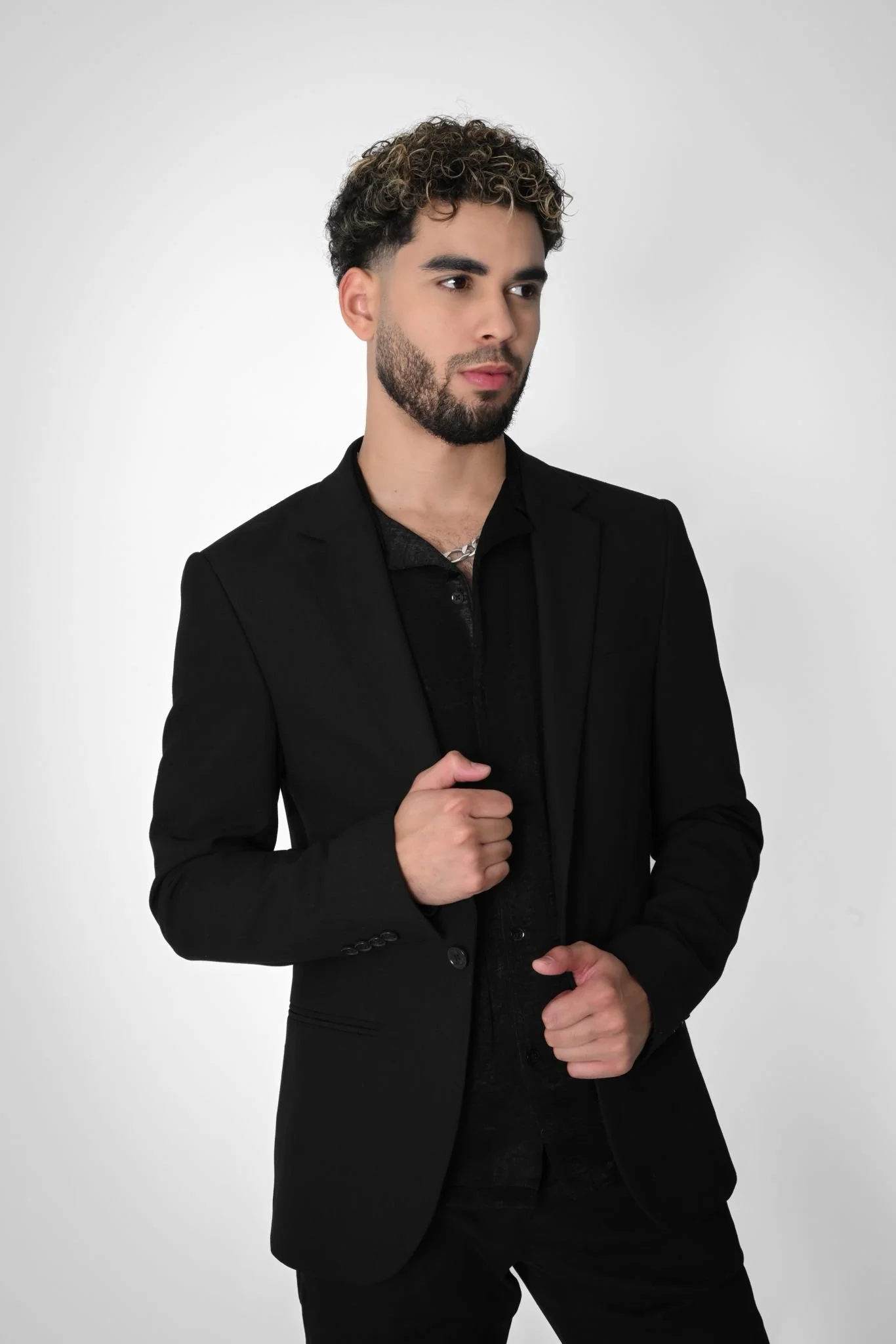 A young man with curly hair and a beard wearing a black blazer and shirt, posing against a plain background.