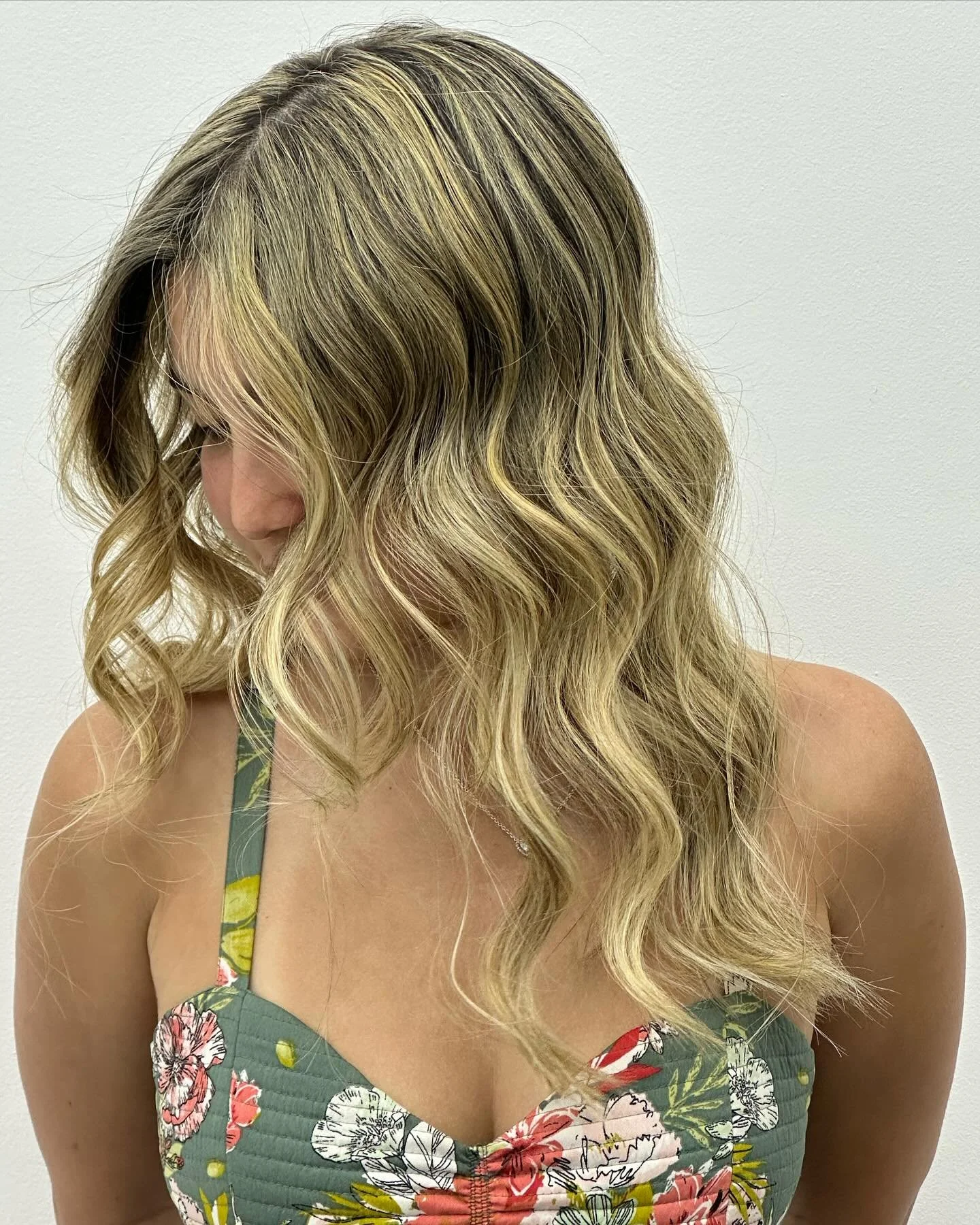 A woman with blonde wavy hair wearing a floral dress, photographed against a plain white wall.