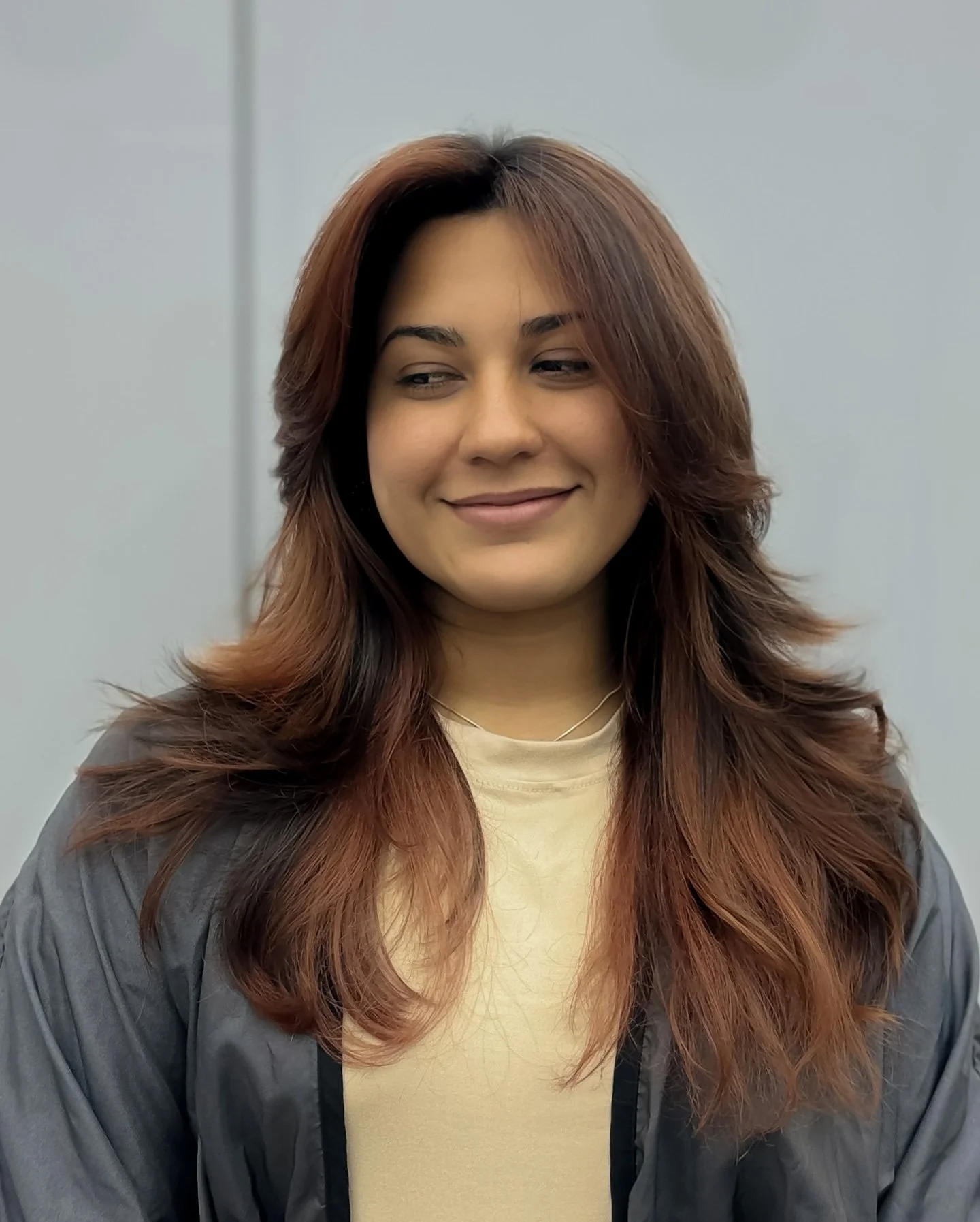 A woman with auburn wavy hair, wearing a gray jacket and a cream-colored top, smiling softly in front of a plain gray wall.