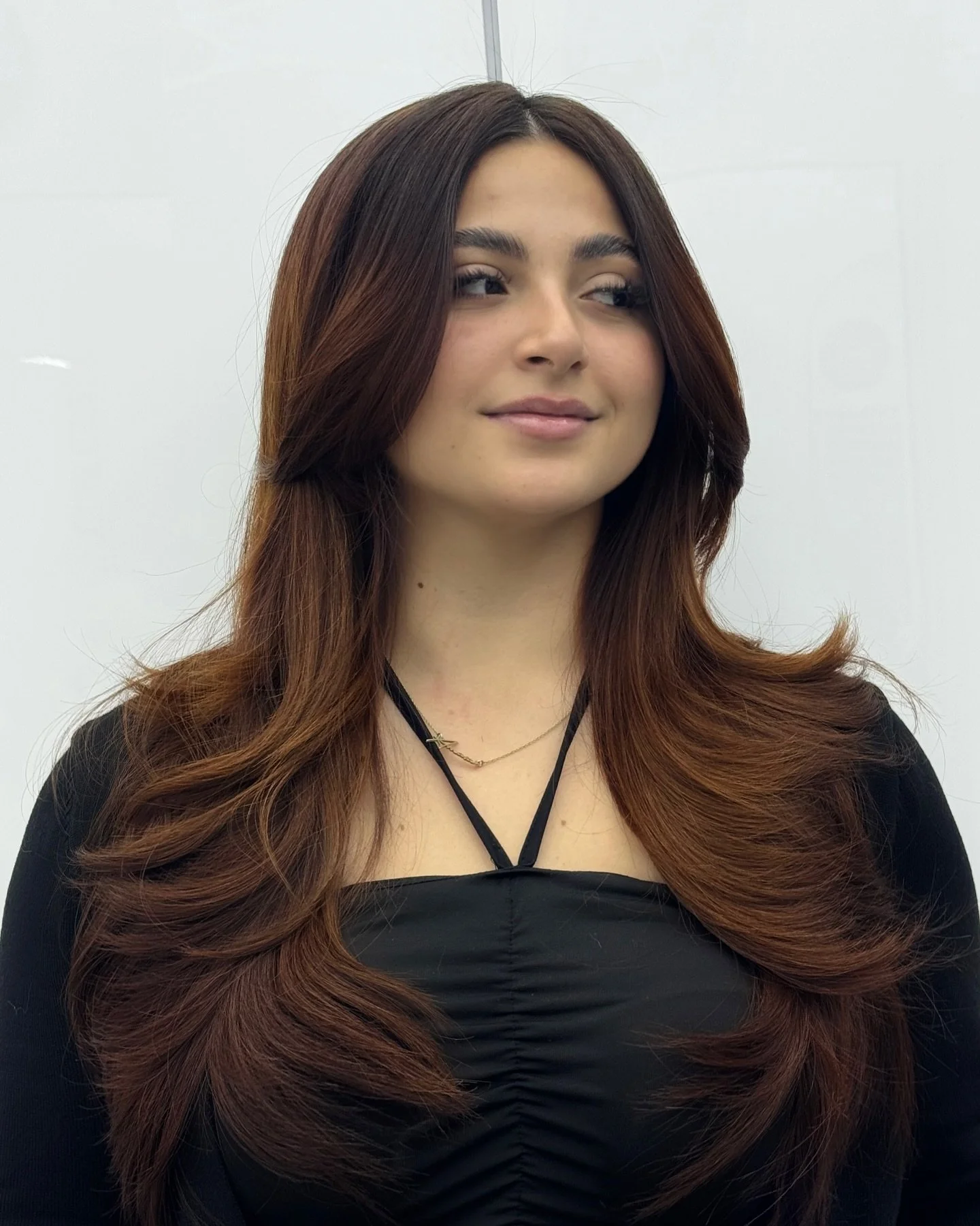 A woman with long, wavy, auburn hair wearing a black top with a beauty mark on her neck, standing against a plain white background.