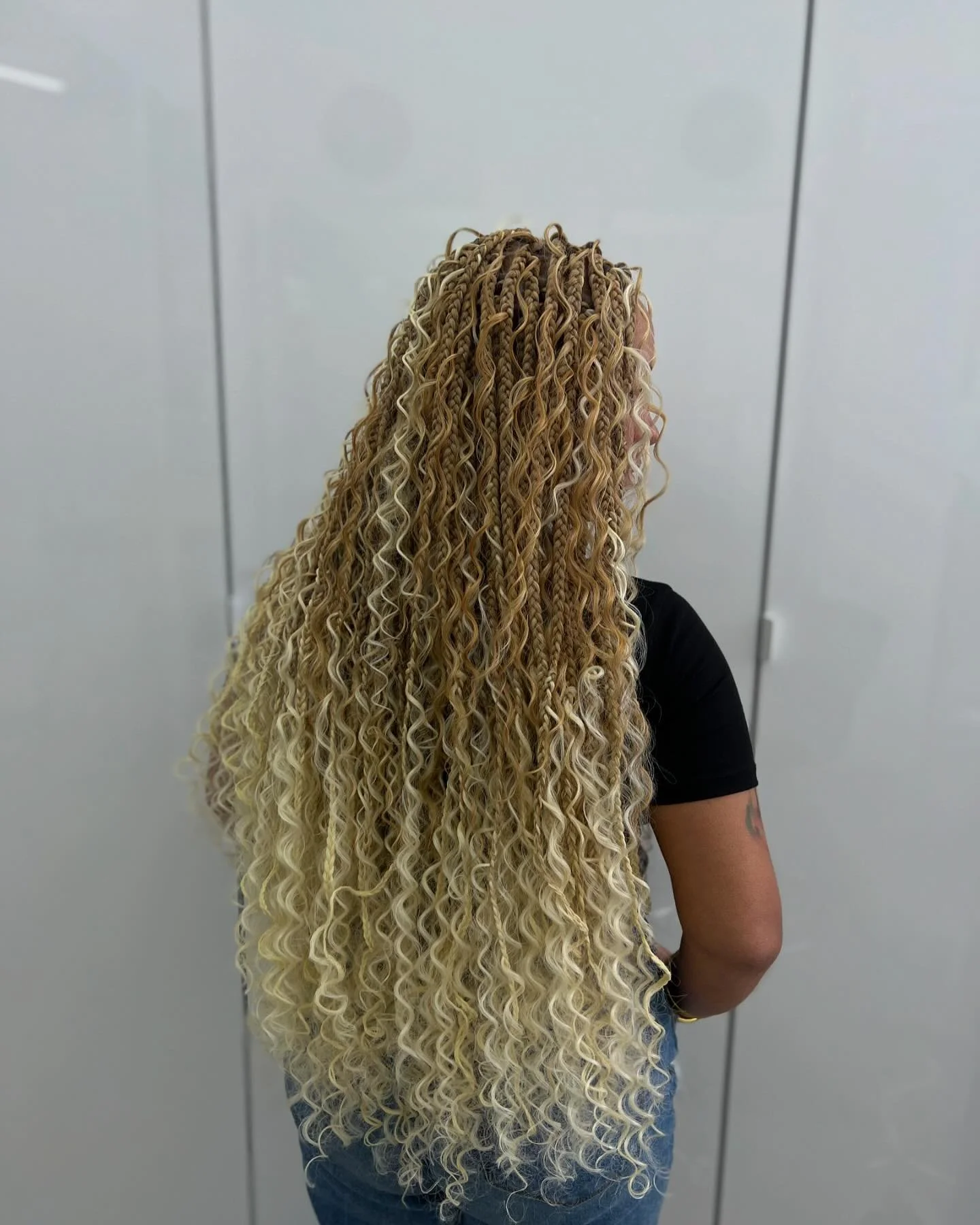 Back view of a person with long, blonde, curly hair standing in front of a white wall with vertical panel lines.