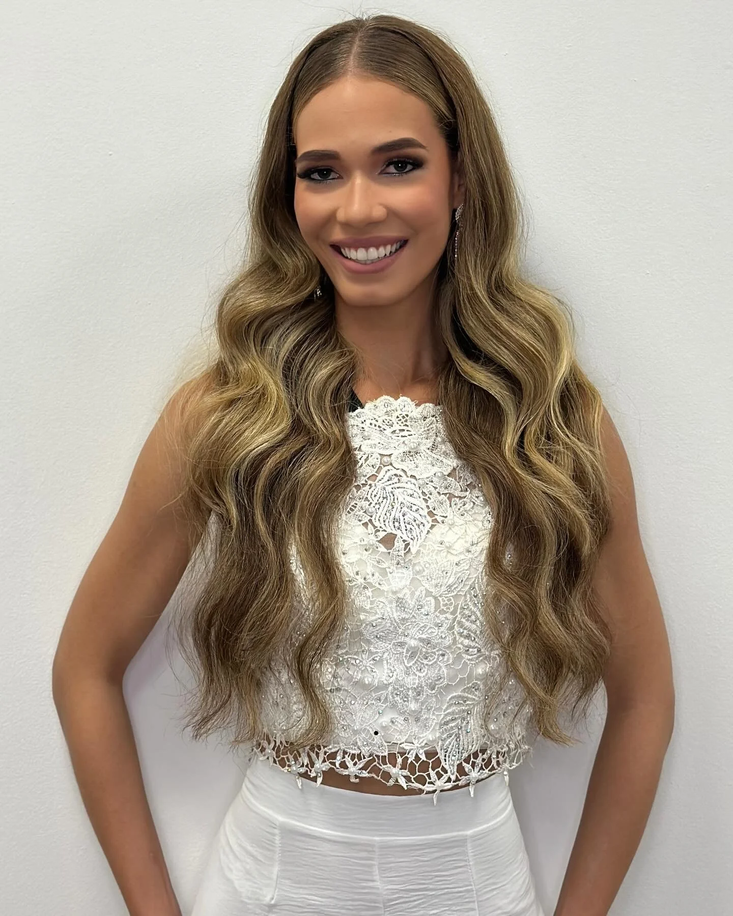 A woman with long wavy hair wearing a white lace sleeveless top and cream skirt, smiling against a plain white wall.