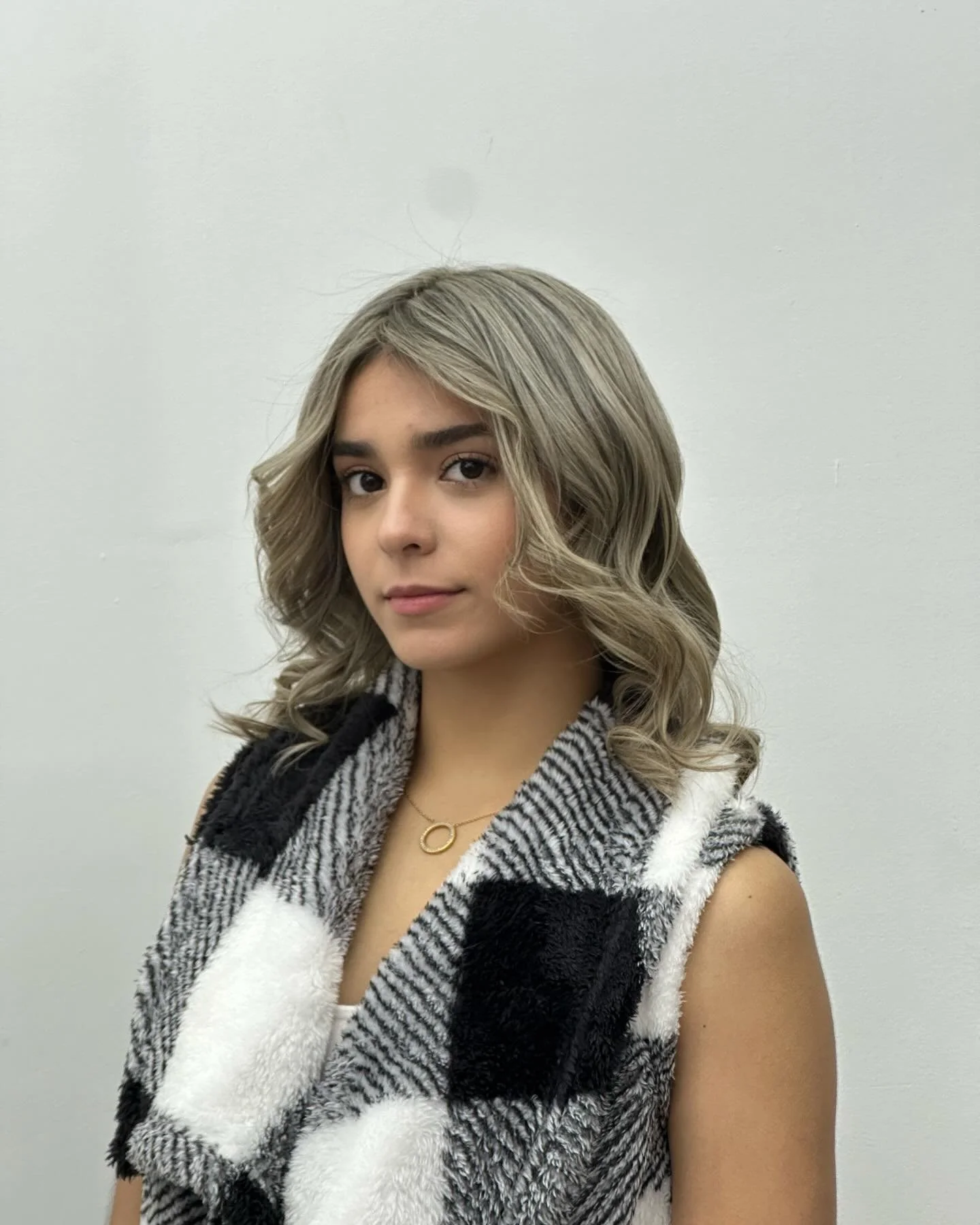 Young woman with wavy blonde hair wearing a black and white animal print vest and a gold necklace, standing against a plain white wall.