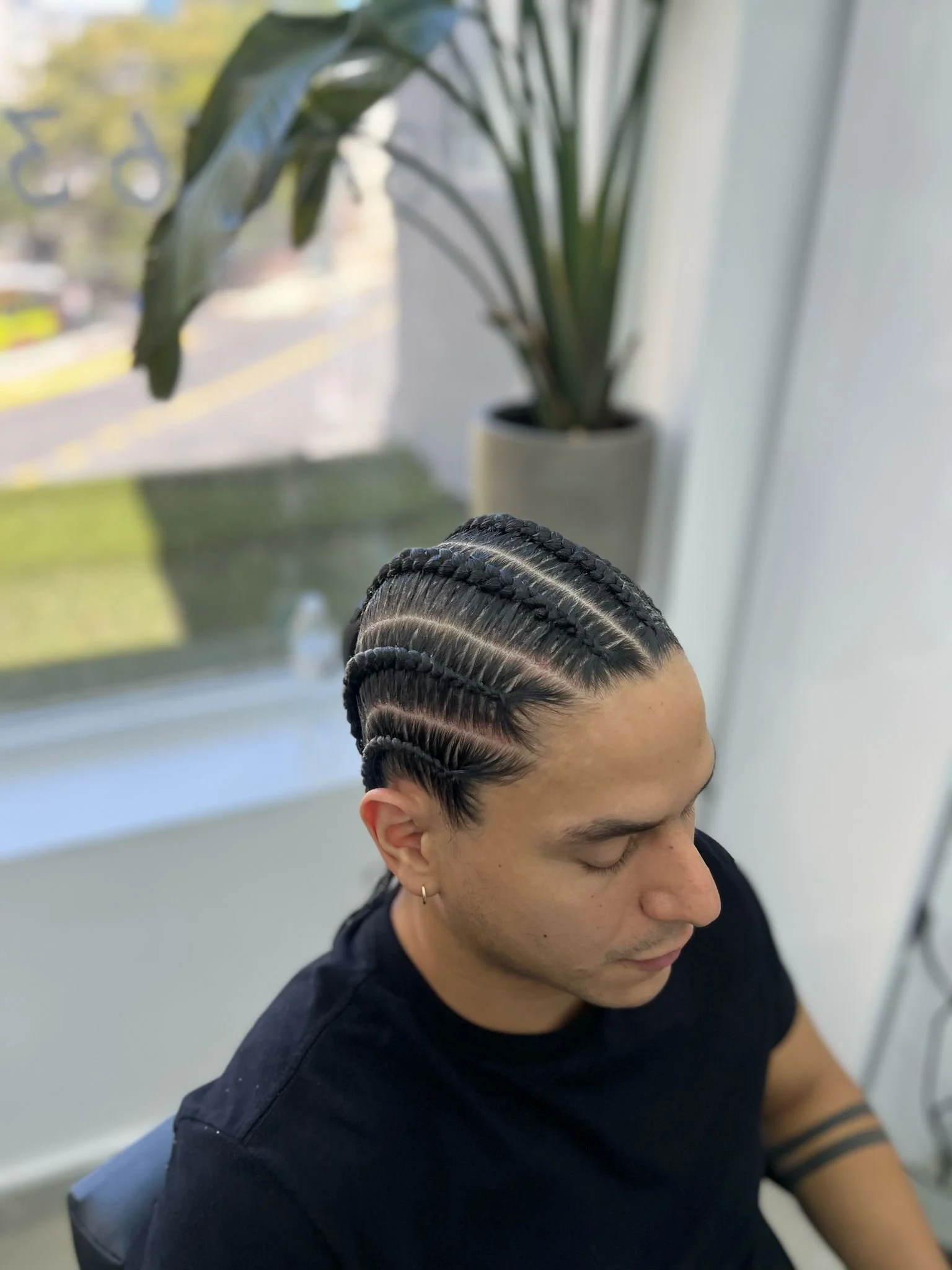 A person with a slicked-back hairstyle featuring multiple tight cornrows sitting indoors near a window, with a plant in the background.