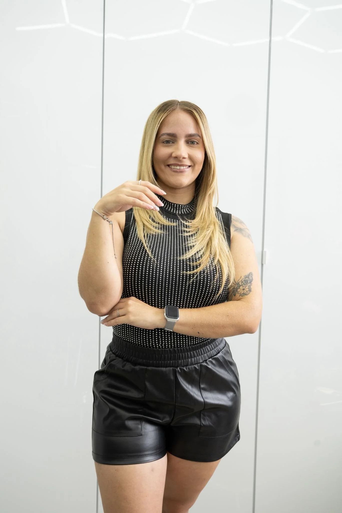A young woman with blonde hair, smiling, wearing a black sleeveless top with white dots, black shorts, and a smartwatch, standing in front of a white background.