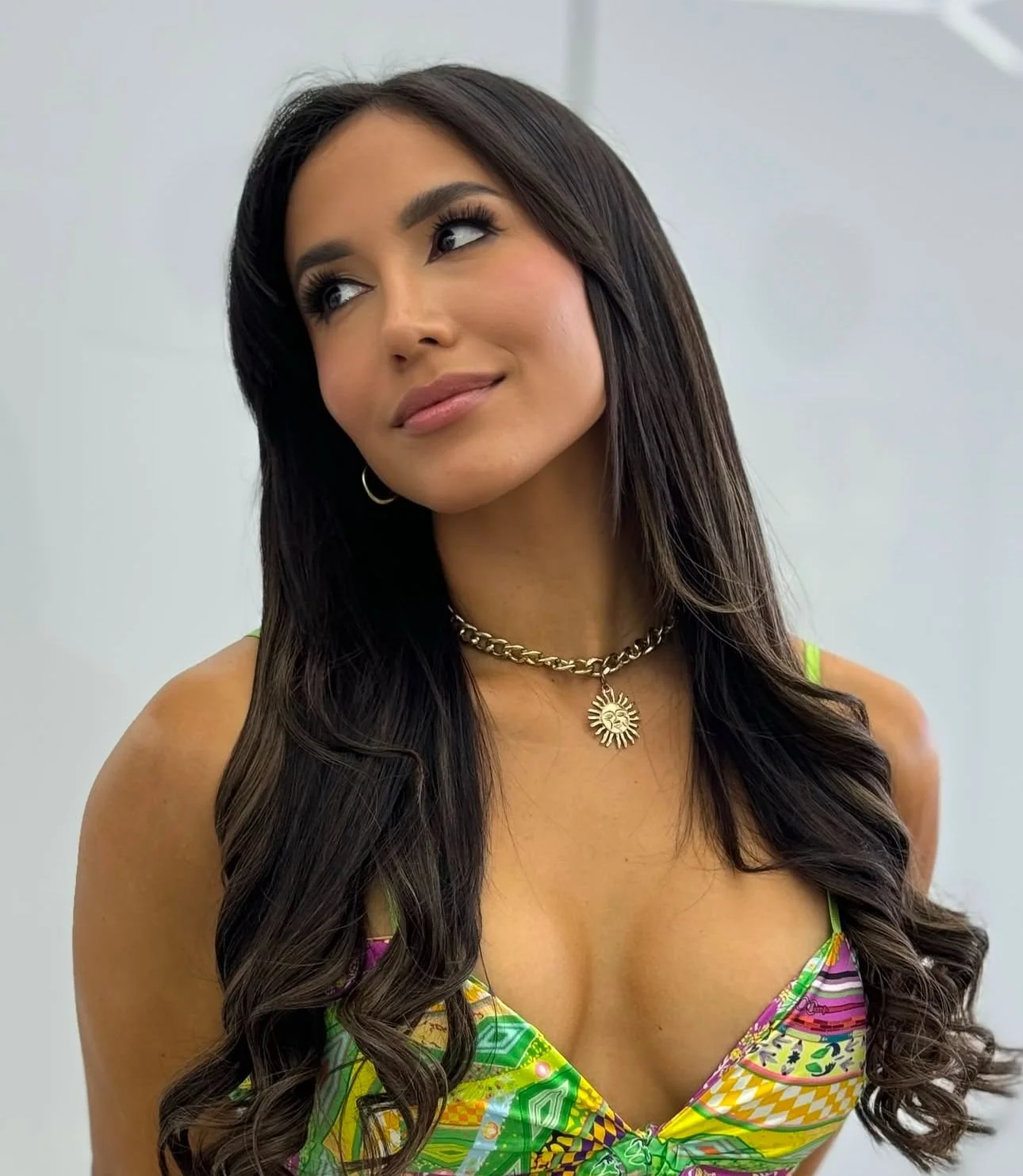 A woman with long wavy dark hair, wearing a colorful patterned dress, gold necklace with a sun pendant, and hoop earrings, looking slightly to her left with a soft smile.