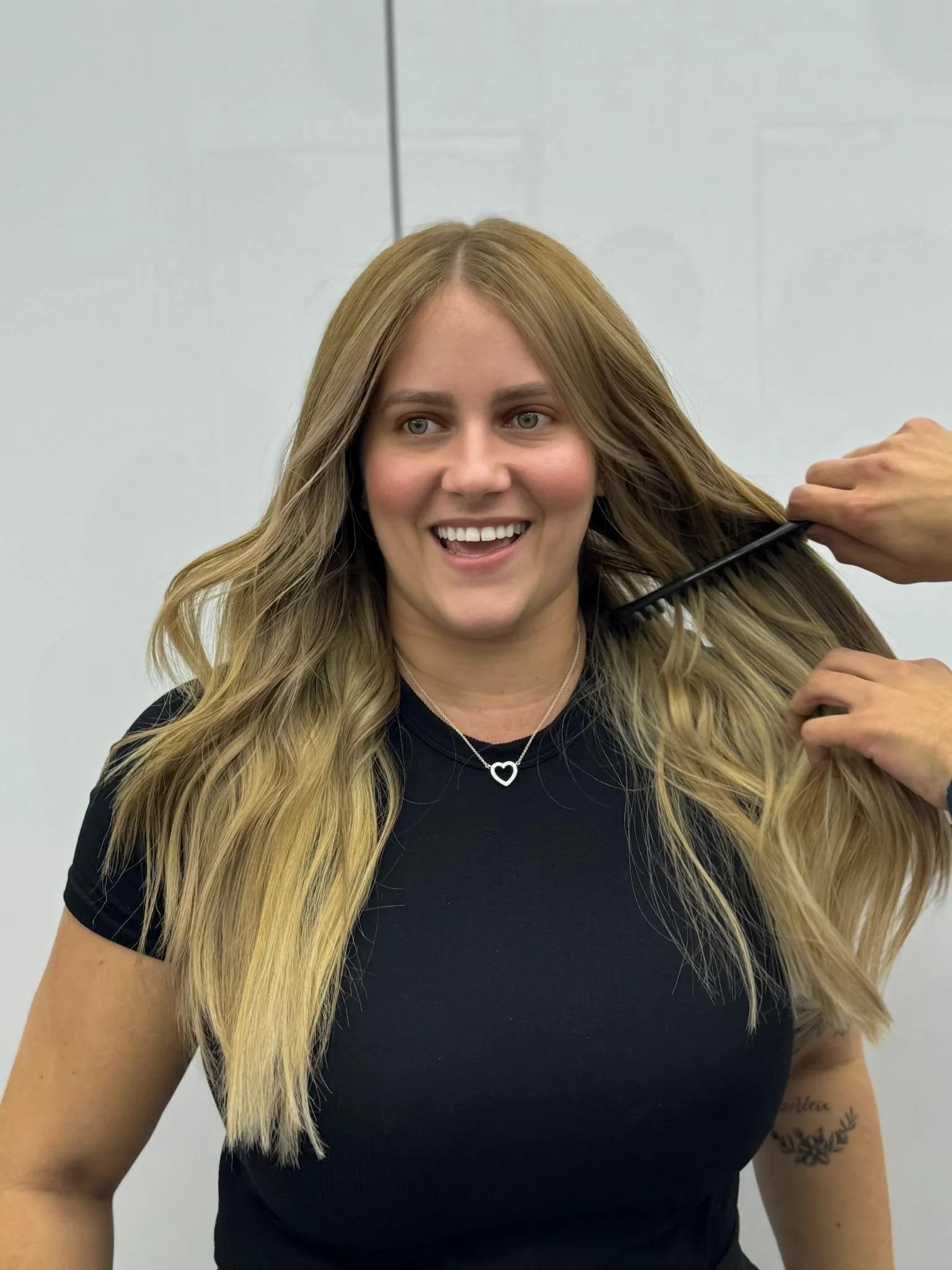 A woman with long, blonde hair smiling and getting her hair styled by a person using a comb.