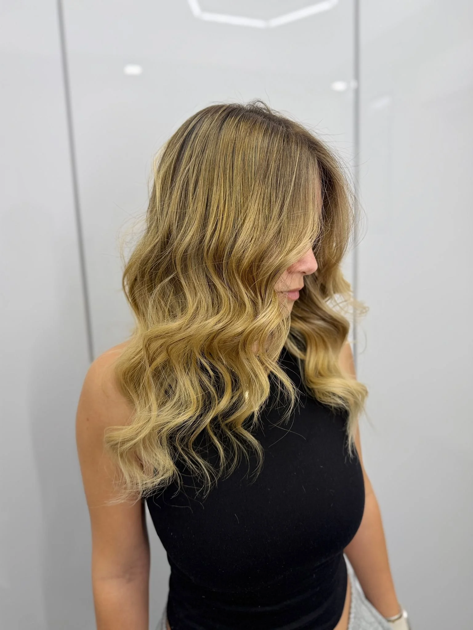 A woman with long, wavy blonde hair styled in loose curls, wearing a black sleeveless top, standing in front of a white background.