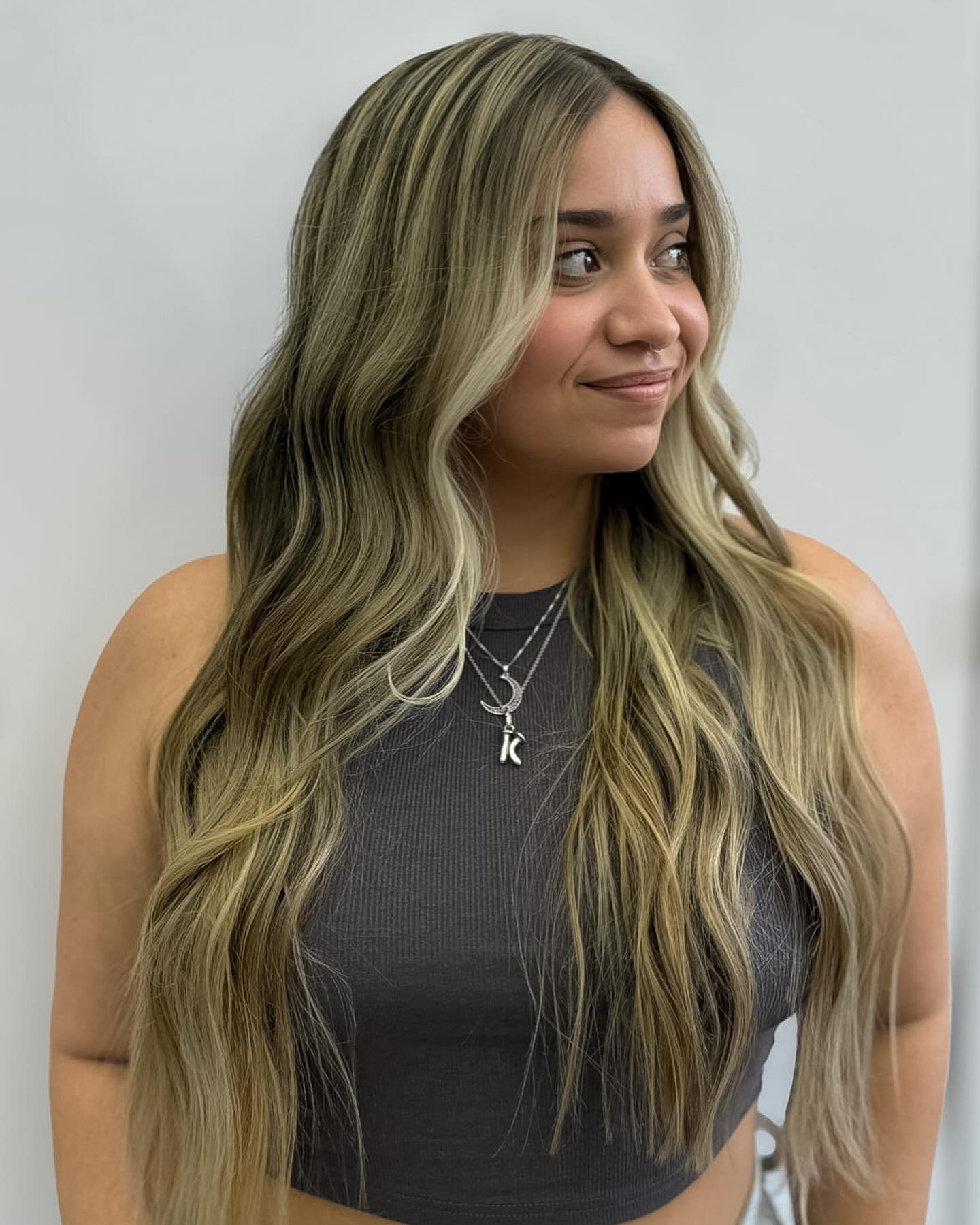 A woman with long, wavy blonde hair wearing a dark gray sleeveless top and layered necklaces with letter and moon charms, smiling and looking to her right against a plain white wall background.