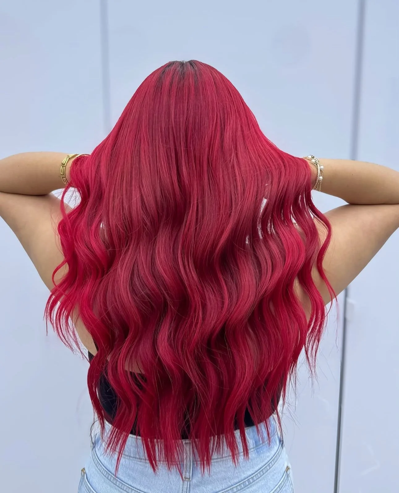 Back view of a woman with long, wavy, bright pink hair, wearing gold jewelry, standing against a light-colored wall.