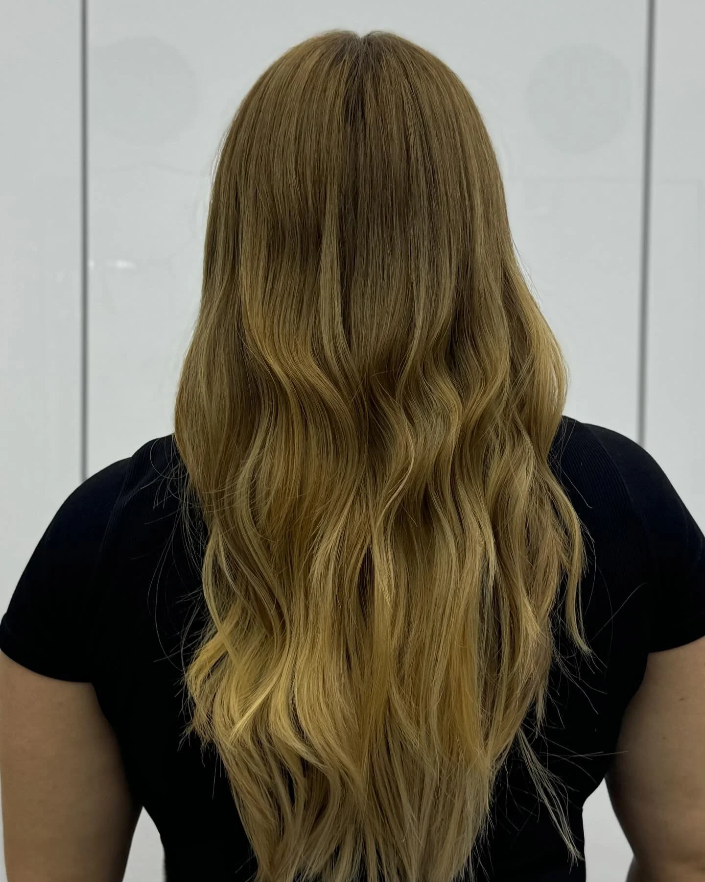 Back view of a woman with long, wavy blonde hair wearing a black top, standing against a white tiled wall.