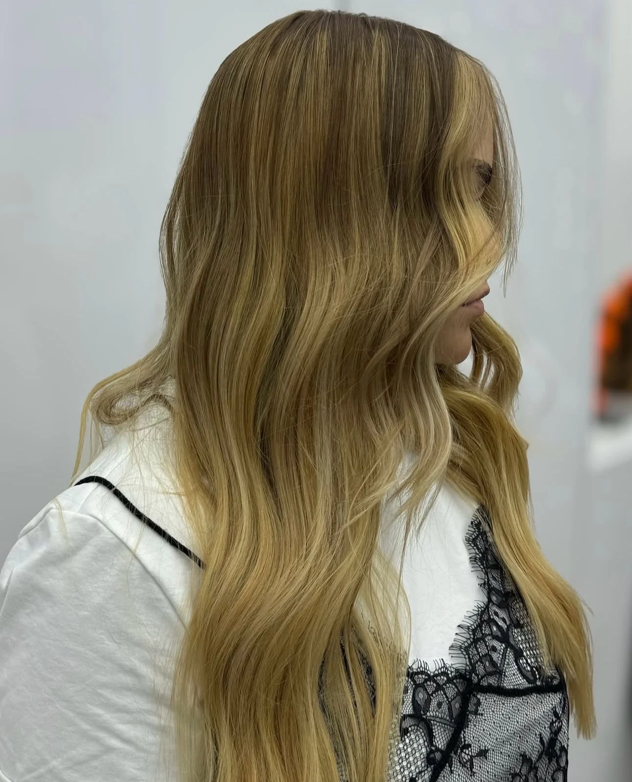 A person with long, wavy blonde hair wearing a white top with black lace detailing, facing to the right against a plain background.