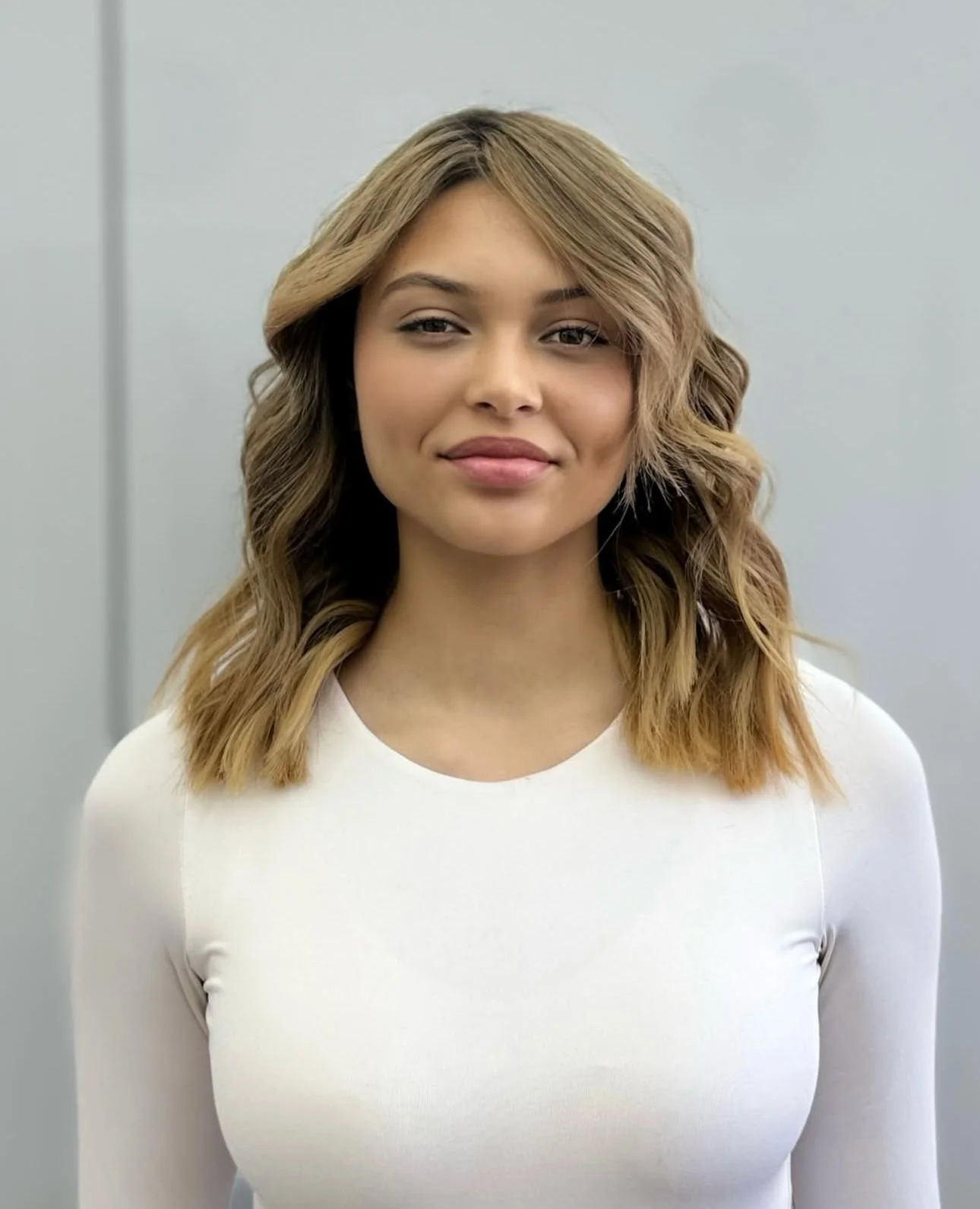 A woman with shoulder-length wavy blond hair, wearing a white top, standing against a plain light gray background.