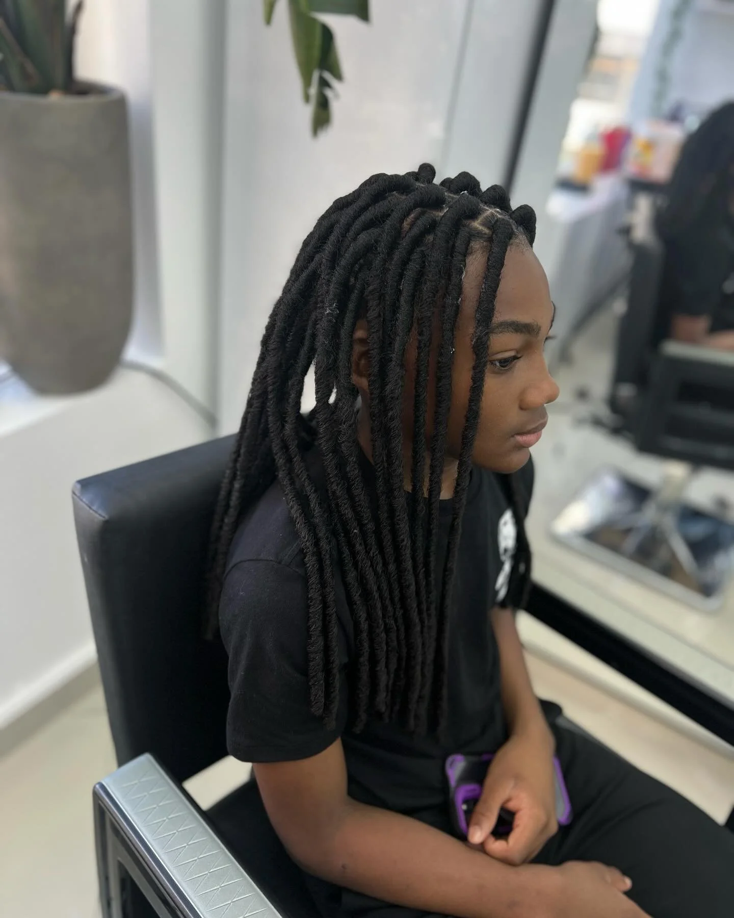 A young person with dreadlocks sitting in a salon chair, looking down, holding a smartphone in their lap, in a salon or barbershop.