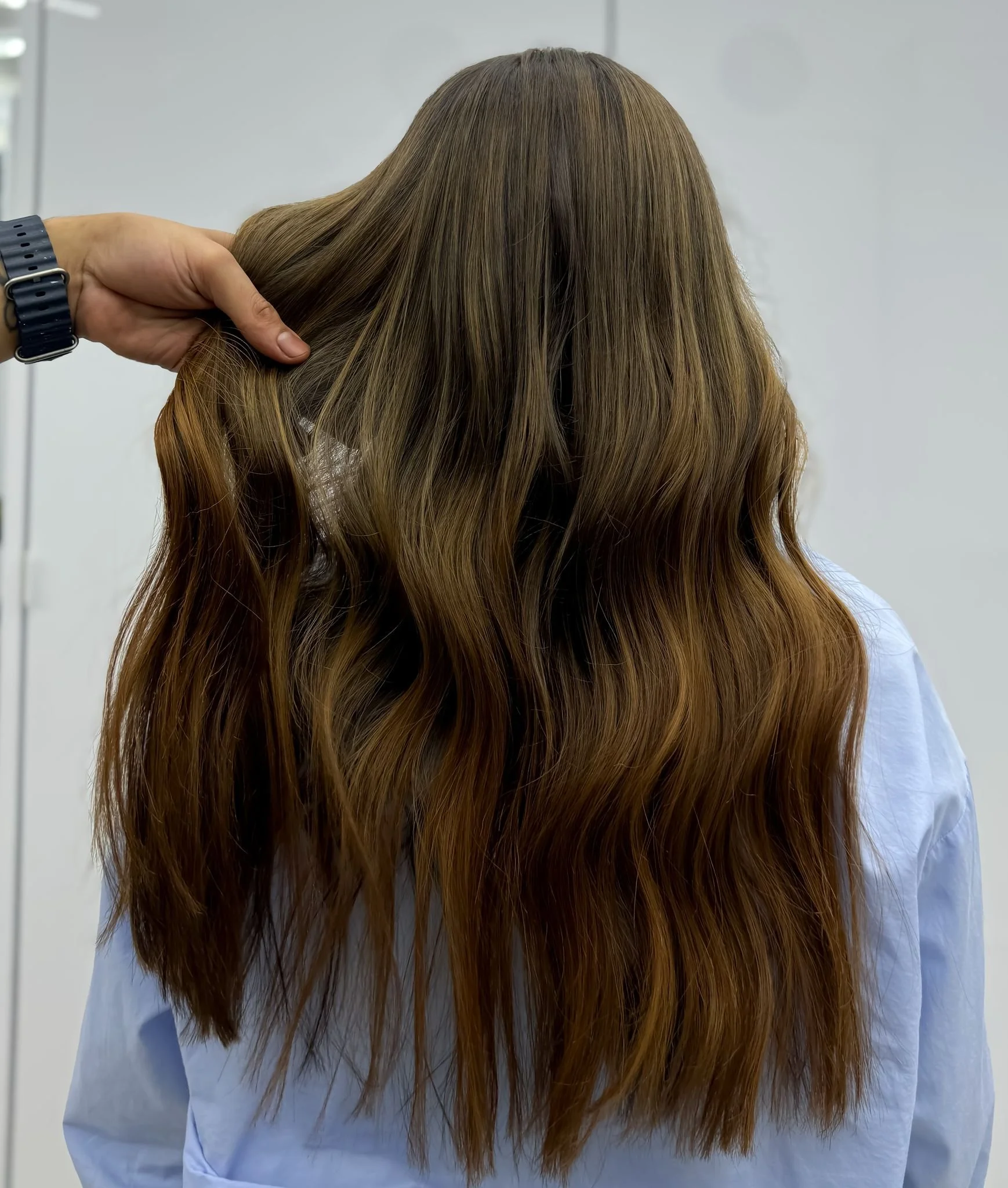 A person with long, wavy brown hair wearing a light blue shirt, viewed from behind, with a hand touching the hair.