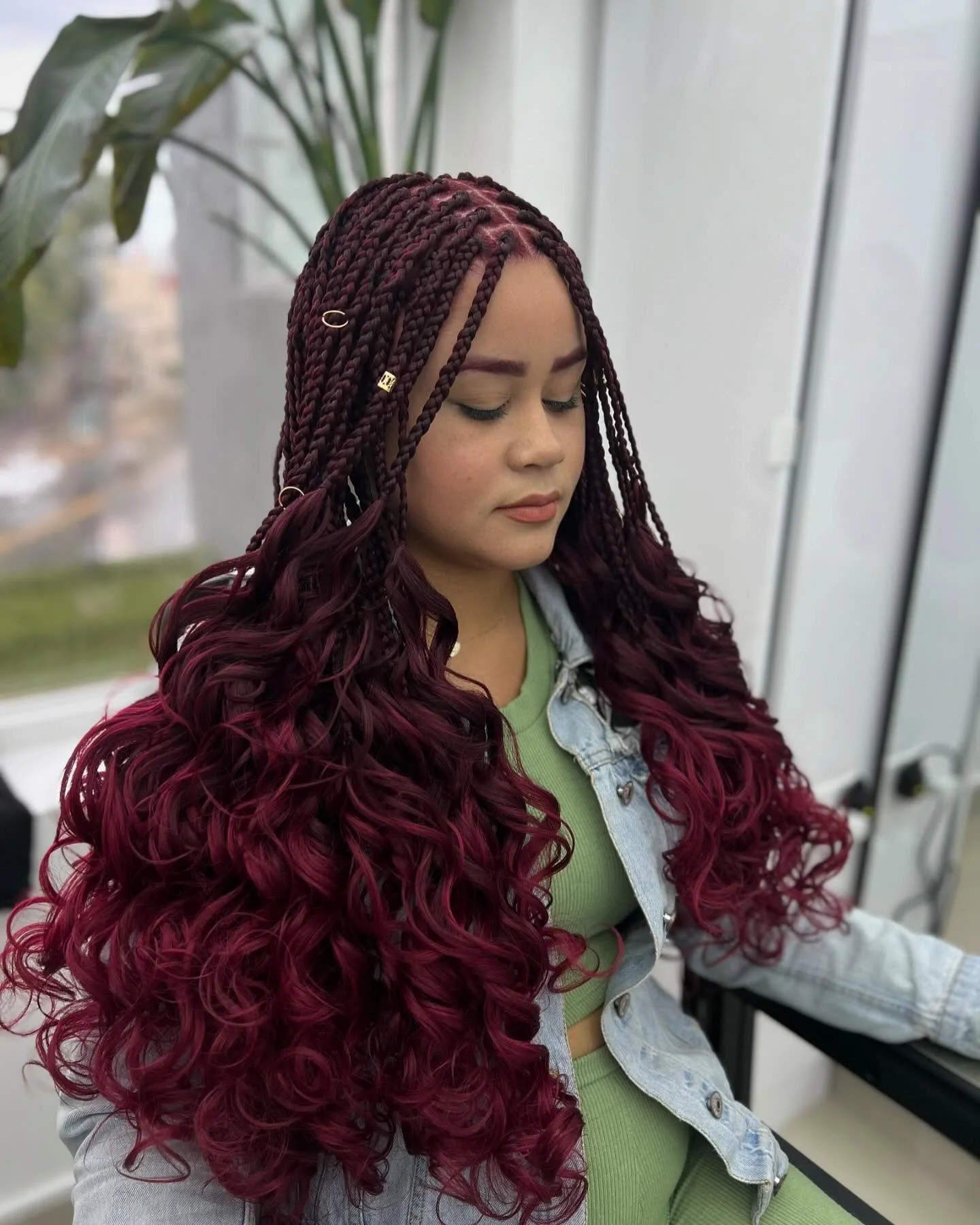 A young woman with long, burgundy curly braids and hair embellishments, sitting on a bus or train with her eyes closed, wearing a light denim jacket and a green top.