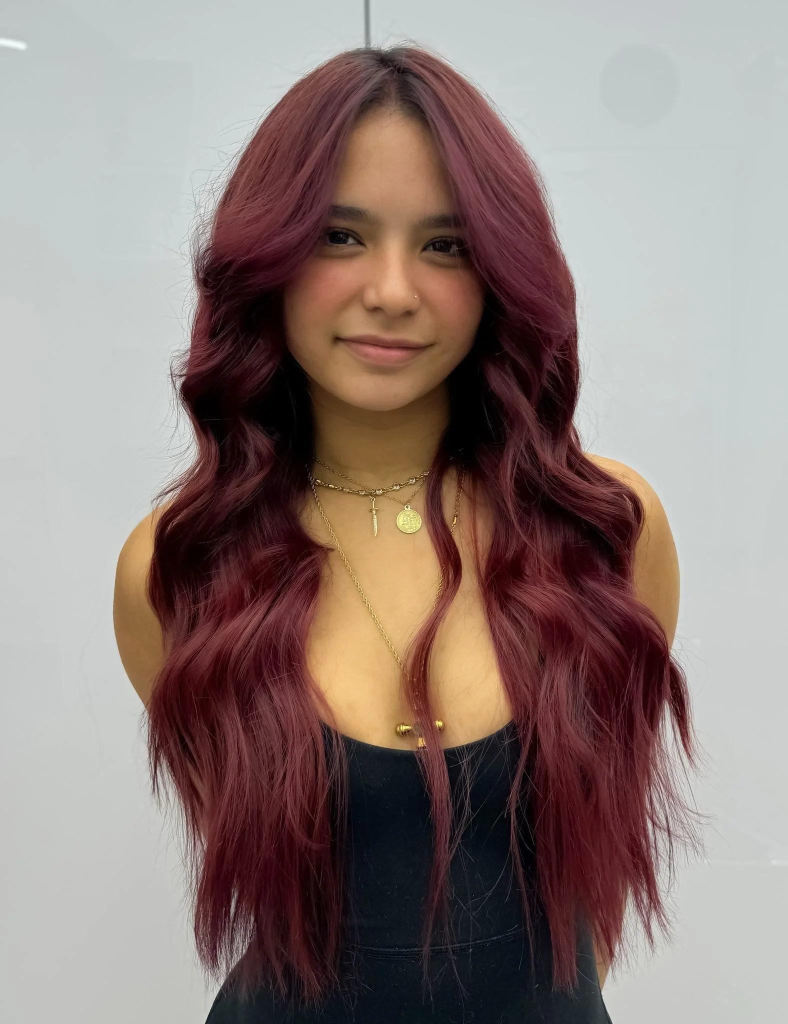 Woman with long, wavy, burgundy hair, wearing multiple layered gold necklaces, standing against a plain light gray background.